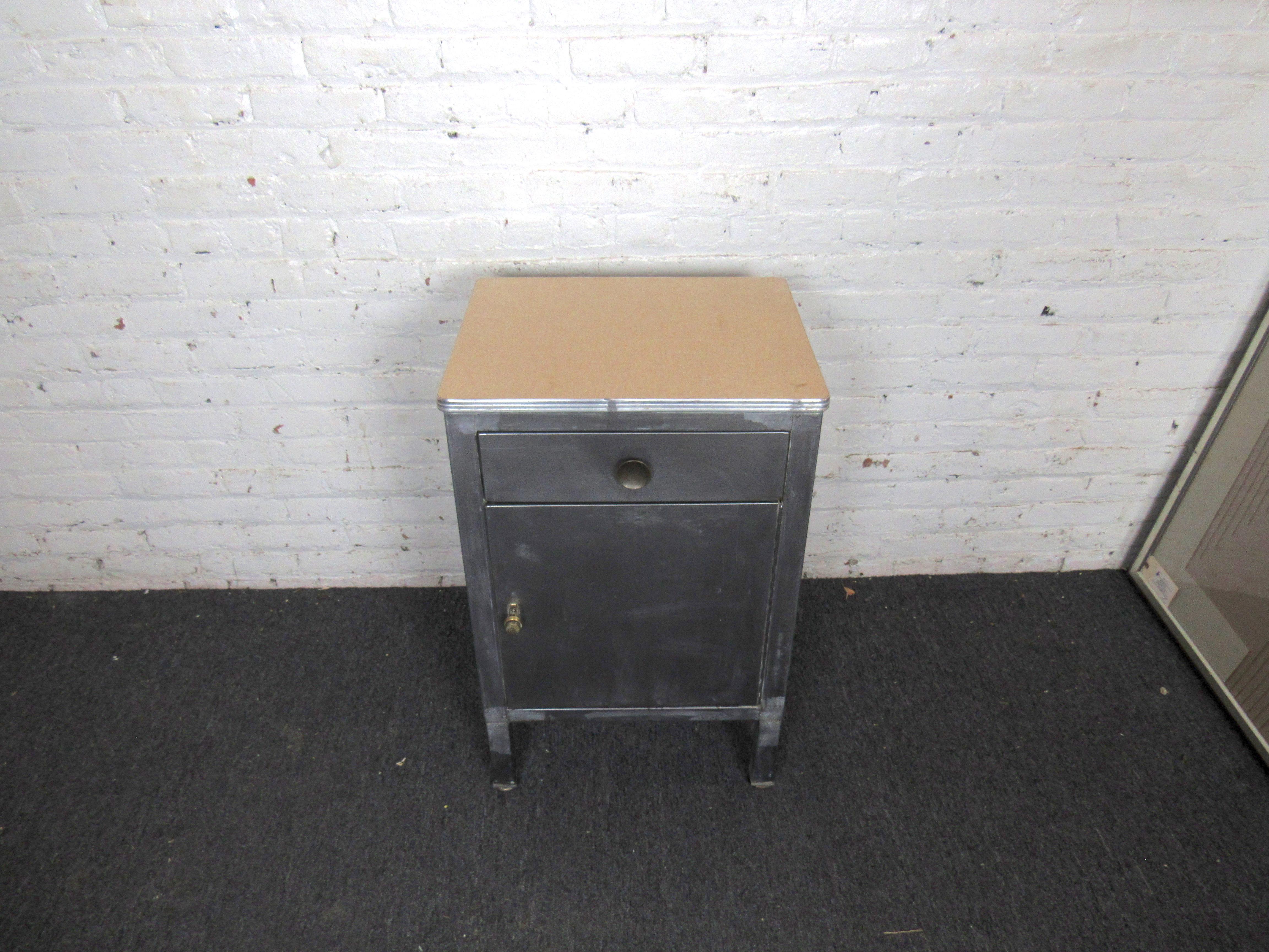 A small, compact and sturdy metal cabinet offering plenty of storage and industrial style. A push button opens the larger door revealing a shelf and large inner compartment, while a top drawer helps organize smaller items. Please confirm item