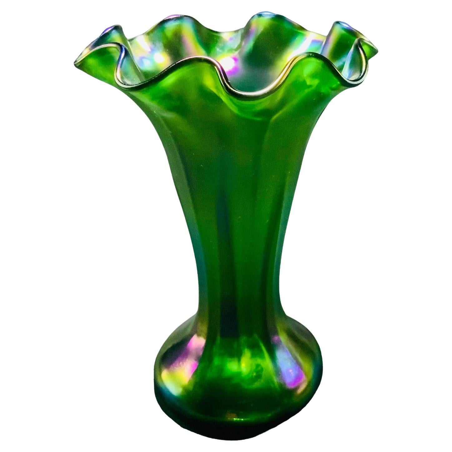 This is a small iridescent Art Glass flower vase. It depicts a different shades of green and few pink, yellow and purple color spots glass flower vase with several ruffles upper border. The whole vase is adorned with reeded bands that match the