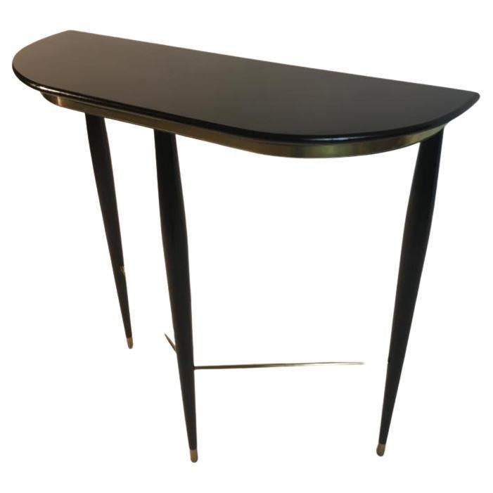 Small Italian console from the 50s, made of black lacquered wood and polished brass. The top is shaped and the legs are tapered towards the bottom. The feet are covered with brass and the very thin legs are connected by a triangle of brass tubes.