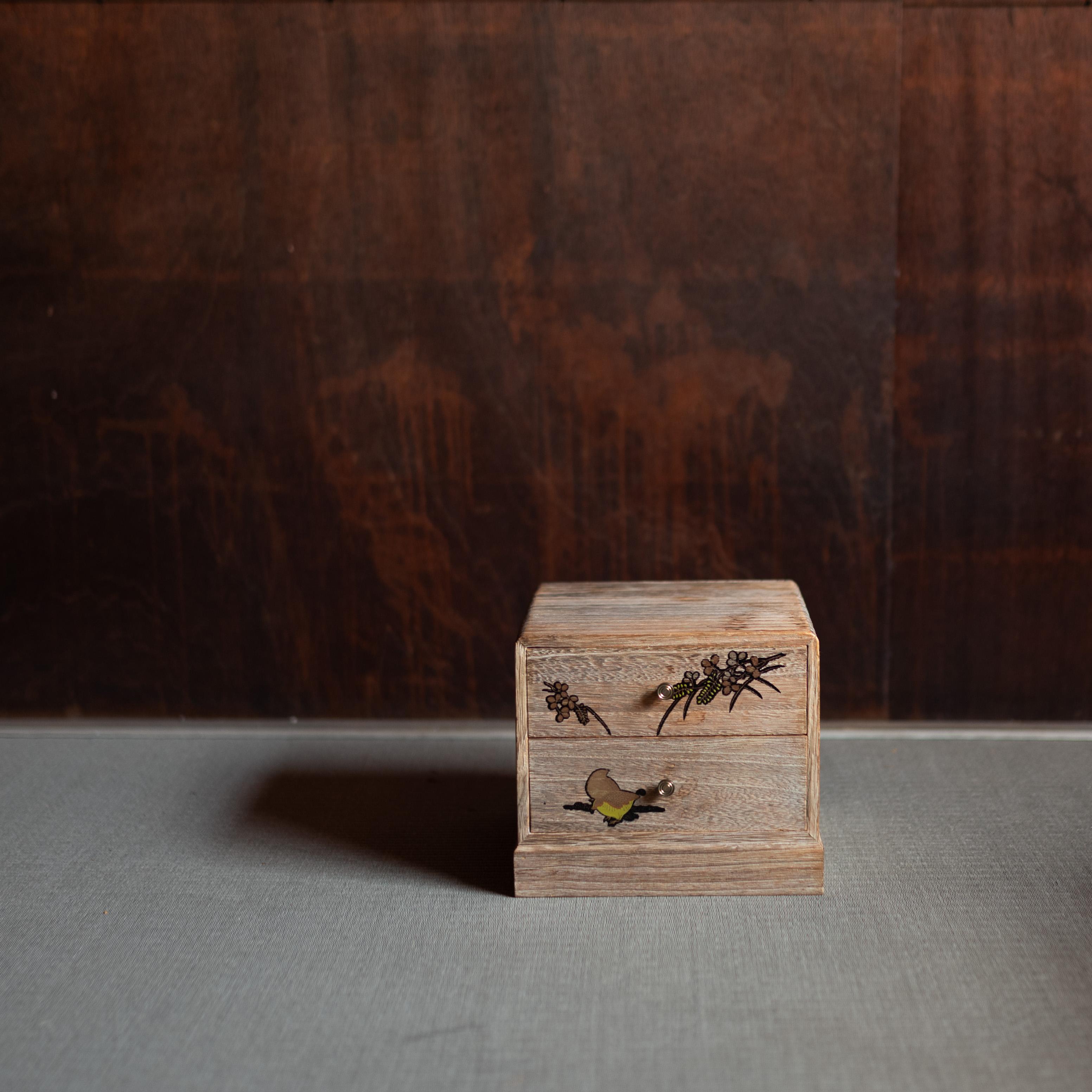 A small Japanese wooden chest of drawers (kodansu), dating from the early 20th century. With two finely made drawers, the front panels are decorated in delicate lacquer work, depicting seasonal flowers and a small bird in flight. The natural wood