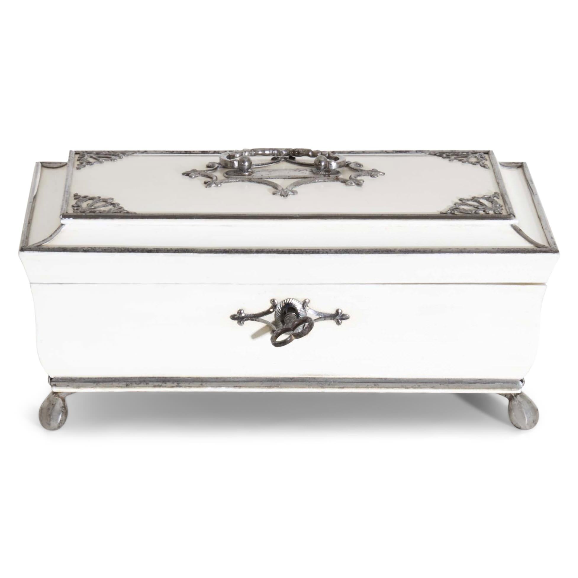 White lidded box standing on small spherical feet with trapezoidal body and lid with a groove. The fittings are silver plated. Key available. Interior veneered in cherry.