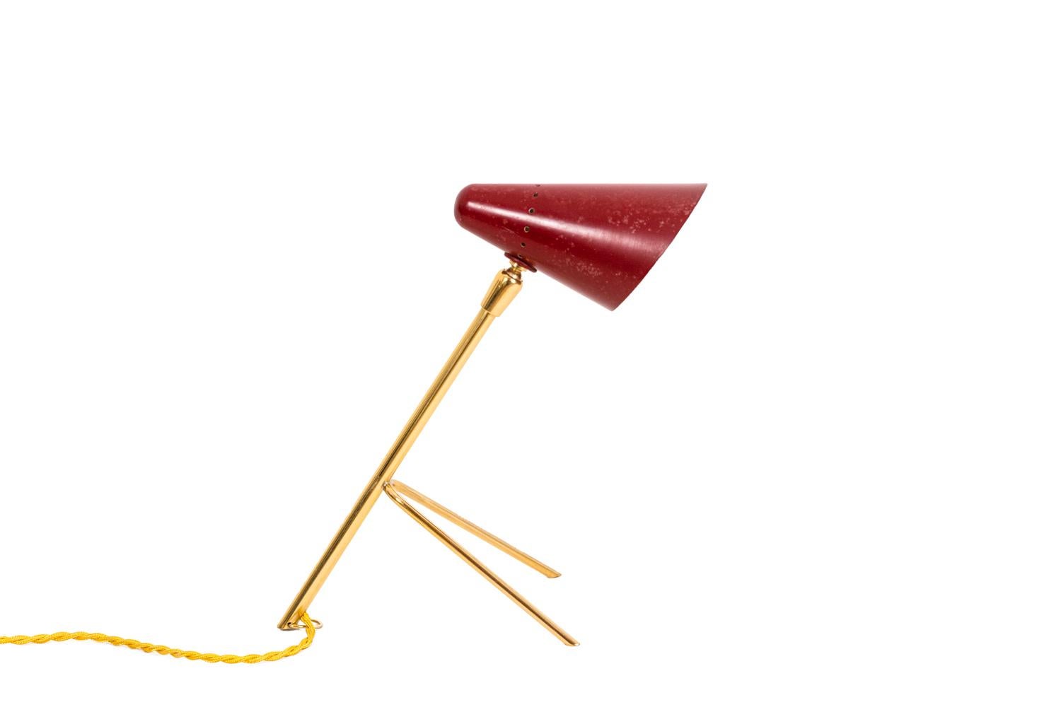 Small tripod lamp standing on three gilt brass legs. Movable conical lampshade in red lacquered metal on the exterior and white lacquered on the interior. Lamp with a hook that permits to hung it like a wall sconce.

Work realized in the 1950s, in