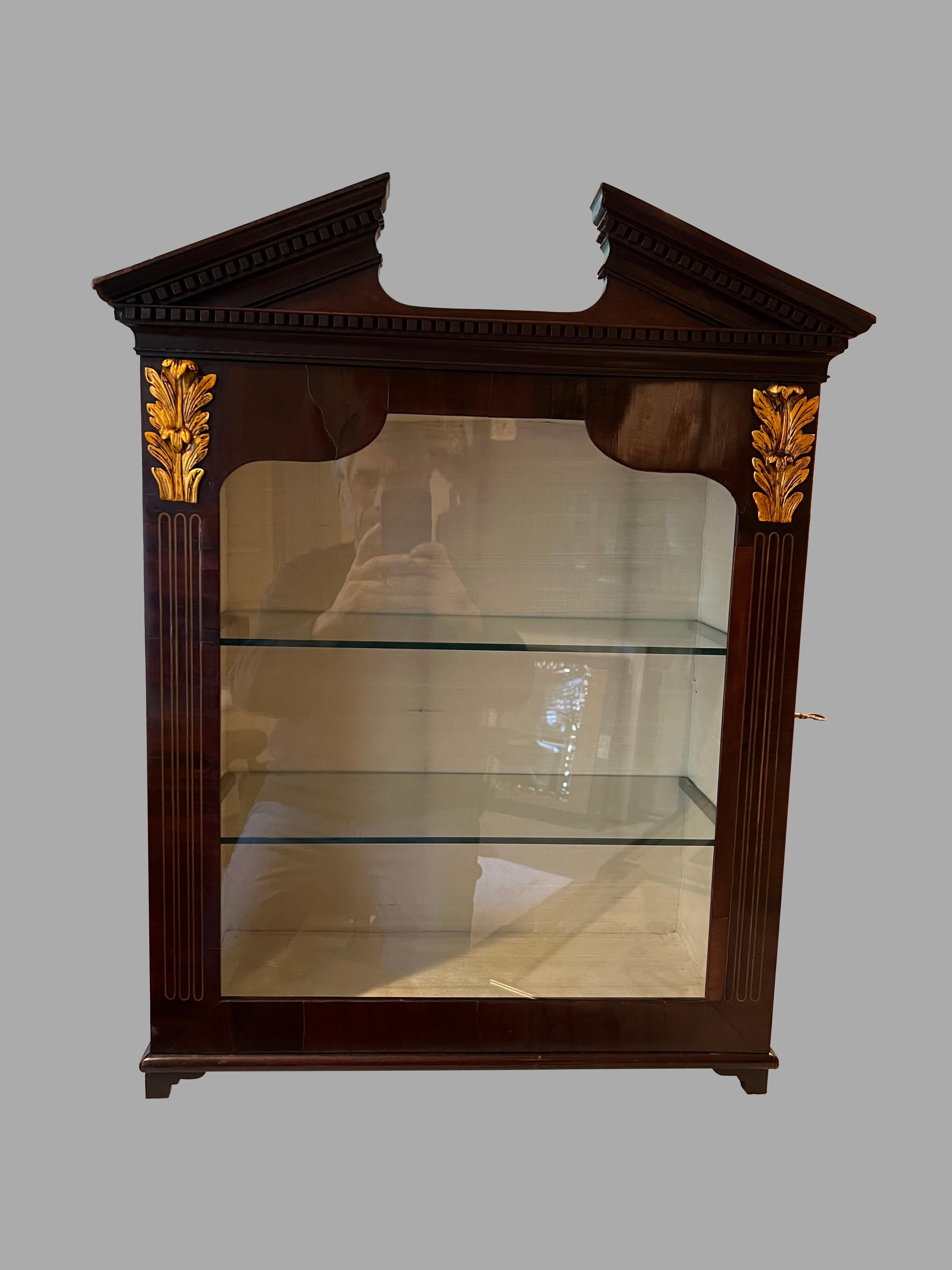 Graziosa vetrina in mogano di epoca tardo-georgiana, il piano a frontone spezzato con modanature a dentelli sopra una coppia di decorazioni dorate a foglie d'acanto e intarsi a linee che incorniciano una porta a vetri. L'interno è foderato in