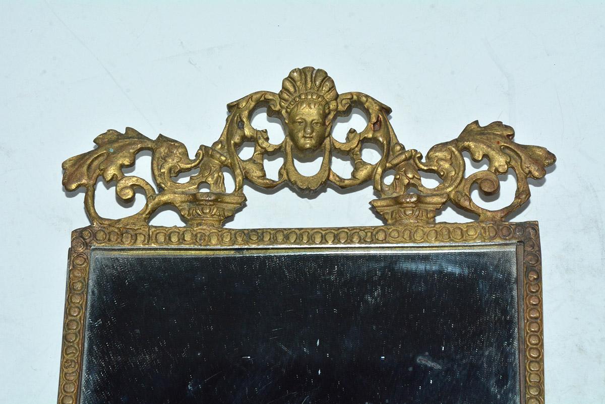 The late Victorian antique ornate mirror has a frame of cast gilt metal. At the top is a mask centered above leaves and vines. At the bottom is a small shield, at the center of additional leaves and vines. The backing is metal as well. The mirror is