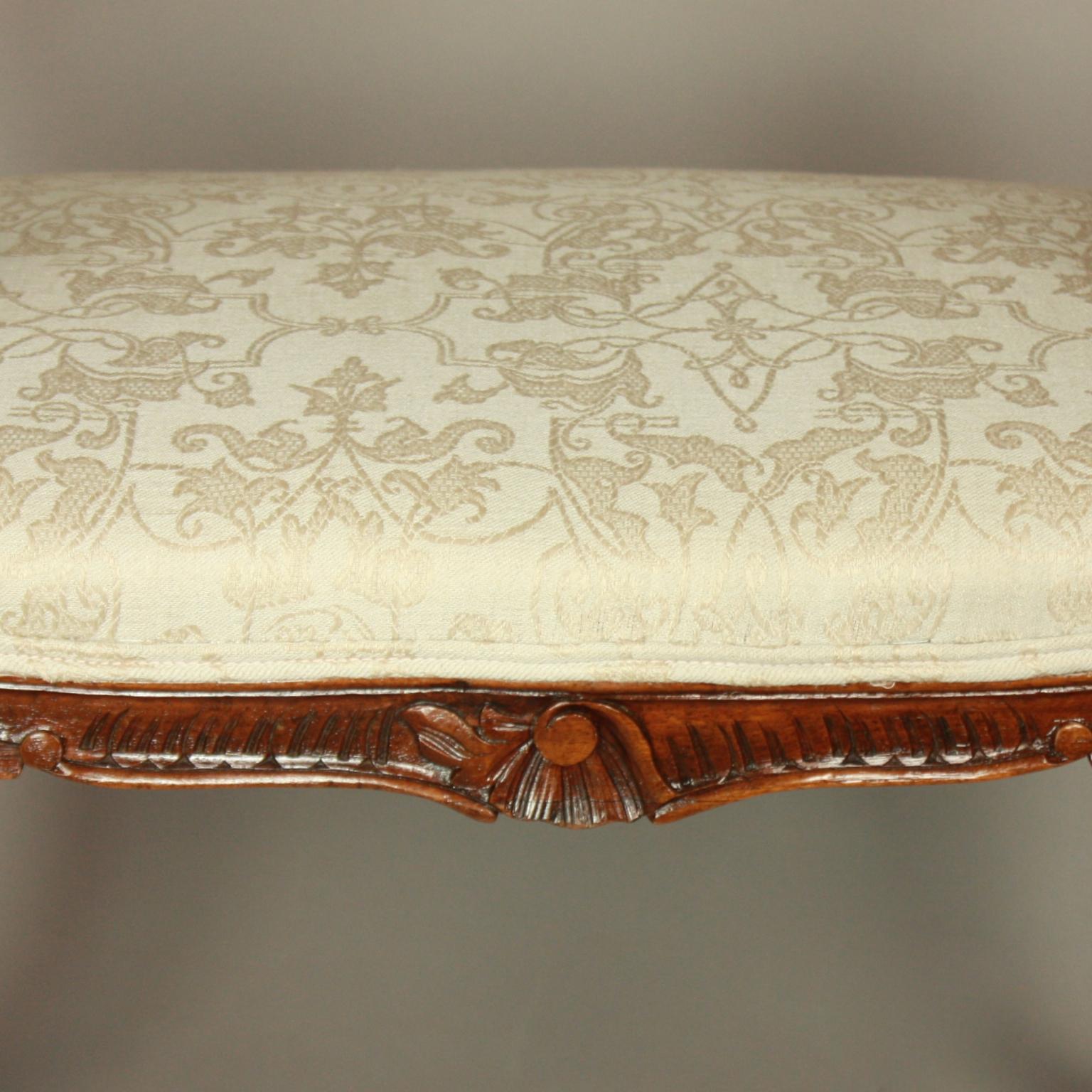 A small and elegant Louis XV banquette or window seat carved in solid walnut featuring scrolled arms and rails, the shaped padded seat above a serpentine frieze carved with rocaille and leaves, the whole supported on finely carved cabriole legs.