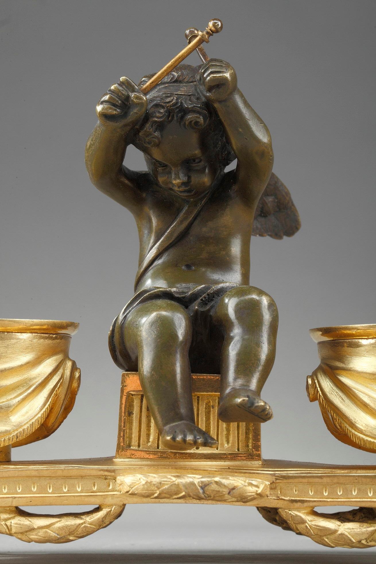 Crafted of gilt and patinated bronze, this small Louis XVI-style inkwell features a winged Cupid playing timbales. He is sitting on a stool, flanked by the two timbales decorated with draperies, hiding the inkwells. The putto is resting on an oval
