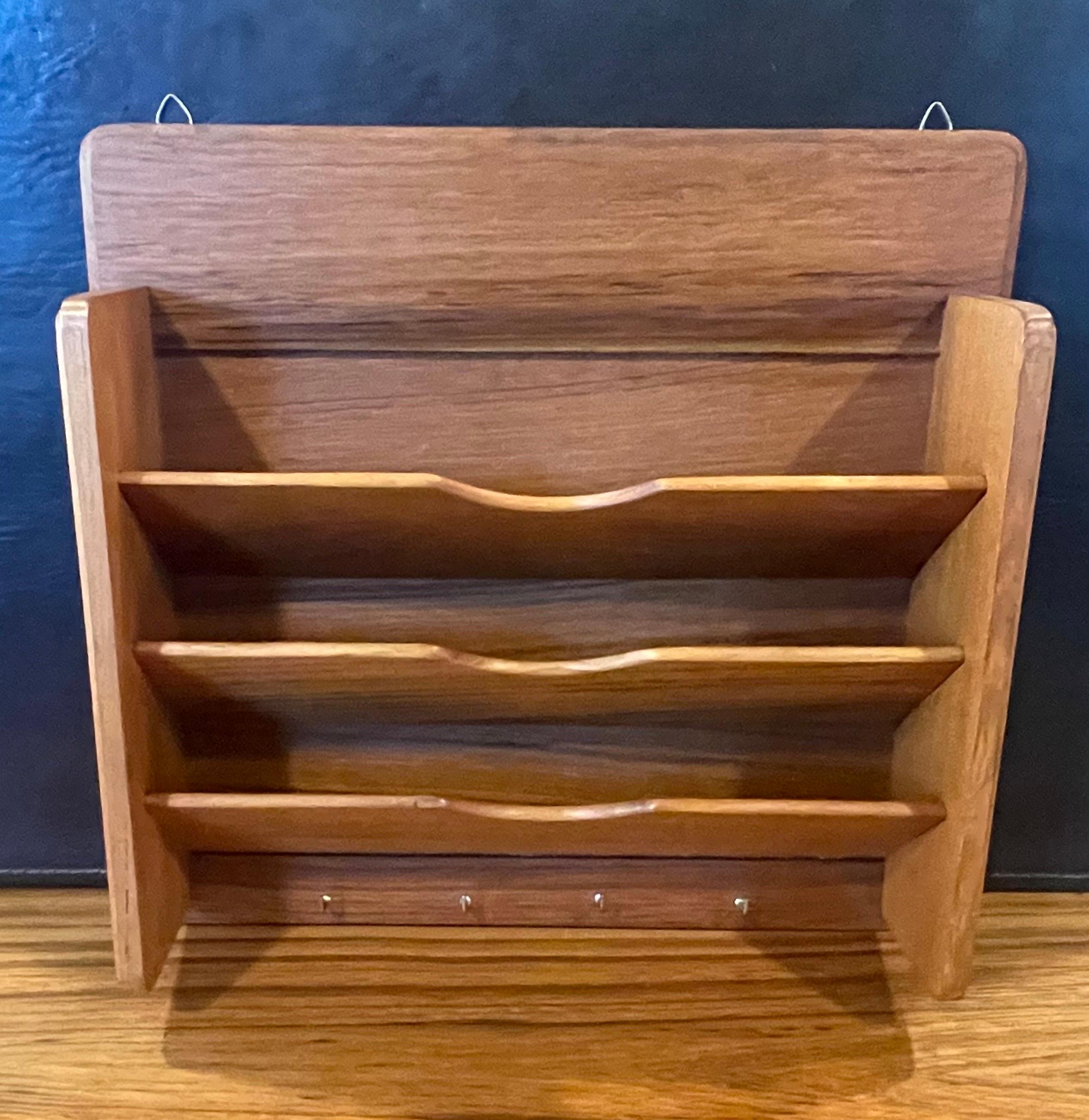 A small MCM teak wall shelf / organizer with three shelves and four brass hooks, circa 1970s. The piece is in very good vintage condition and measures 12.375