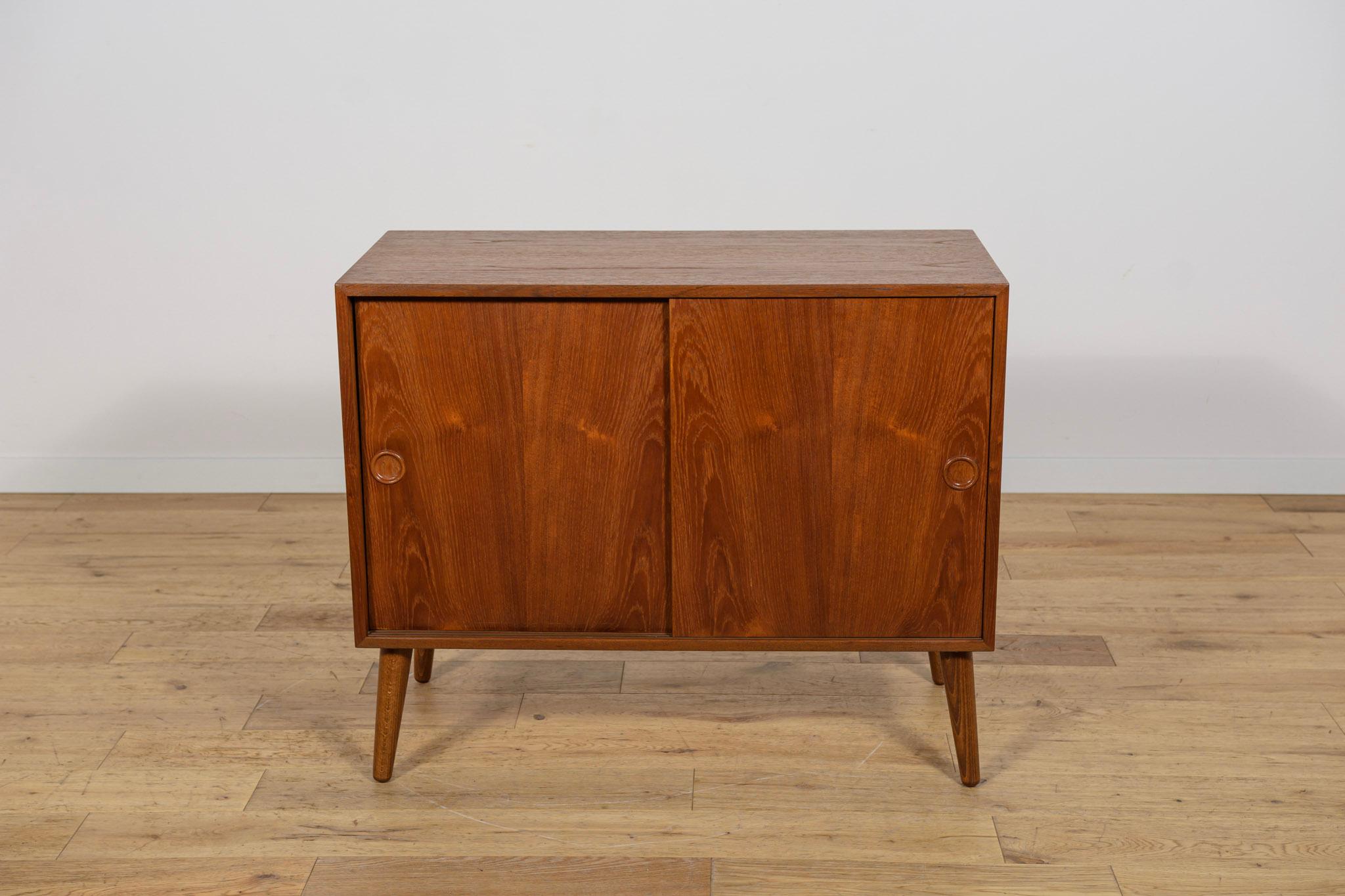 Este pequeño aparador de madera de teca de mediados de siglo fue fabricado en Dinamarca por Feldballes Møbelfabrik en la década de 1960. Tiene dos puertas correderas.  En el interior de la cómoda hay un estante y dos cajones. La cómoda tiene