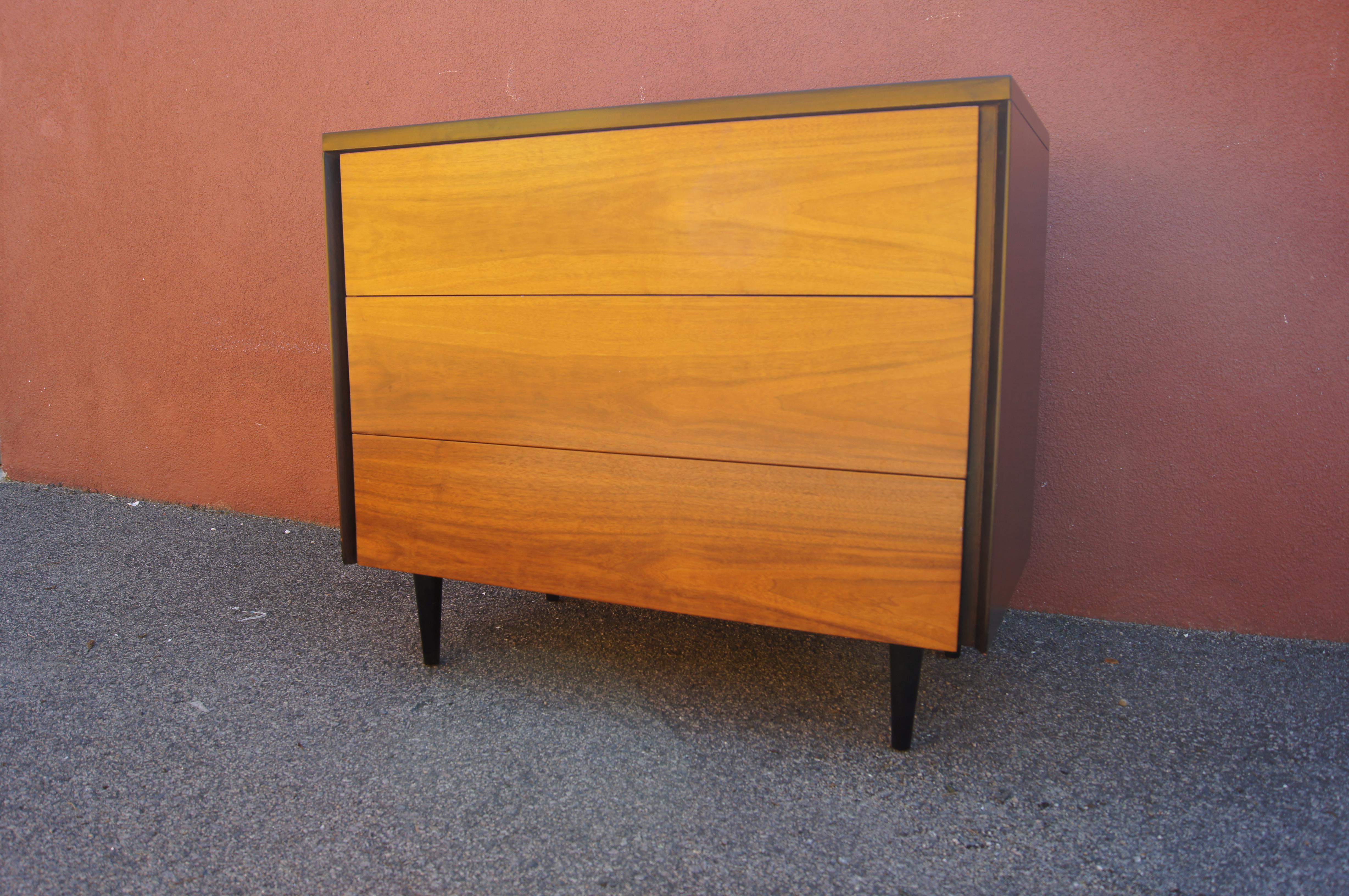 This midcentury three-drawer dresser features a walnut case on tapered black-painted metal legs. The drawer are recessed within the frame and their integral pulls reached at the sides, creating a clean front.

No maker's mark.