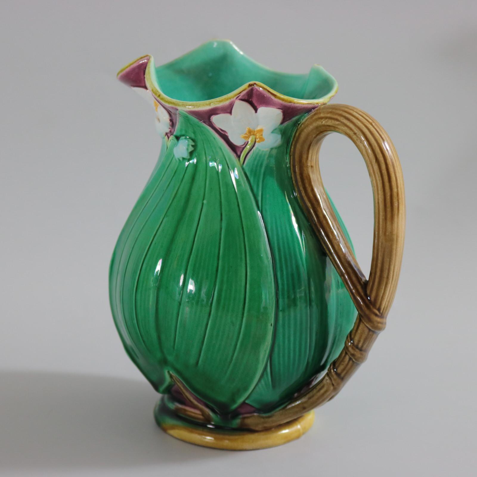 Minton Majolica jug/pitcher which features flowering lilies and lily pads. Colouration: green, dark pink, white, are predominant. The piece bears maker's marks for the Minton pottery. Bears a pattern number, '1228'. Marks include a factory specific