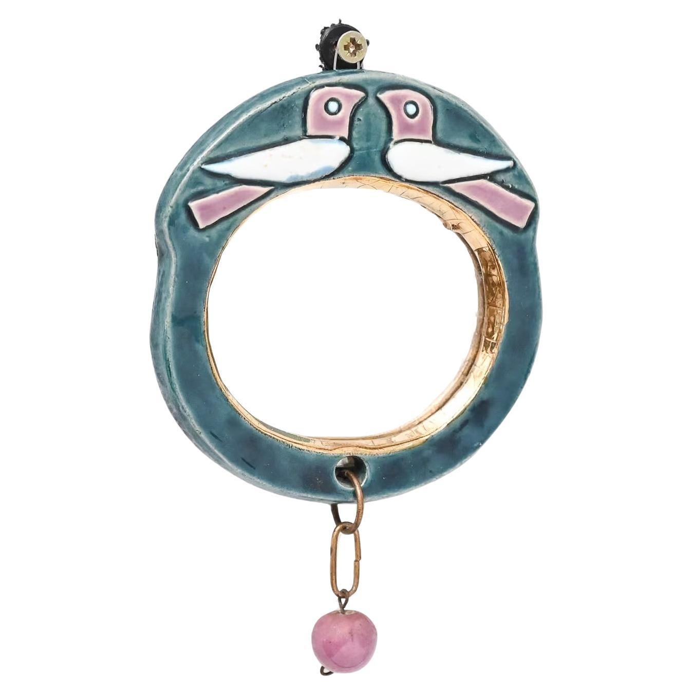 Petit miroir de poche circulaire en céramique de Whiting, décoré dans les tons bleu vert, blanc, rose et or. Surmontée d'une paire d'oiseaux en relief, une courte chaîne en laiton avec une perle rose est attachée à la base. 

Mithé Artistics était