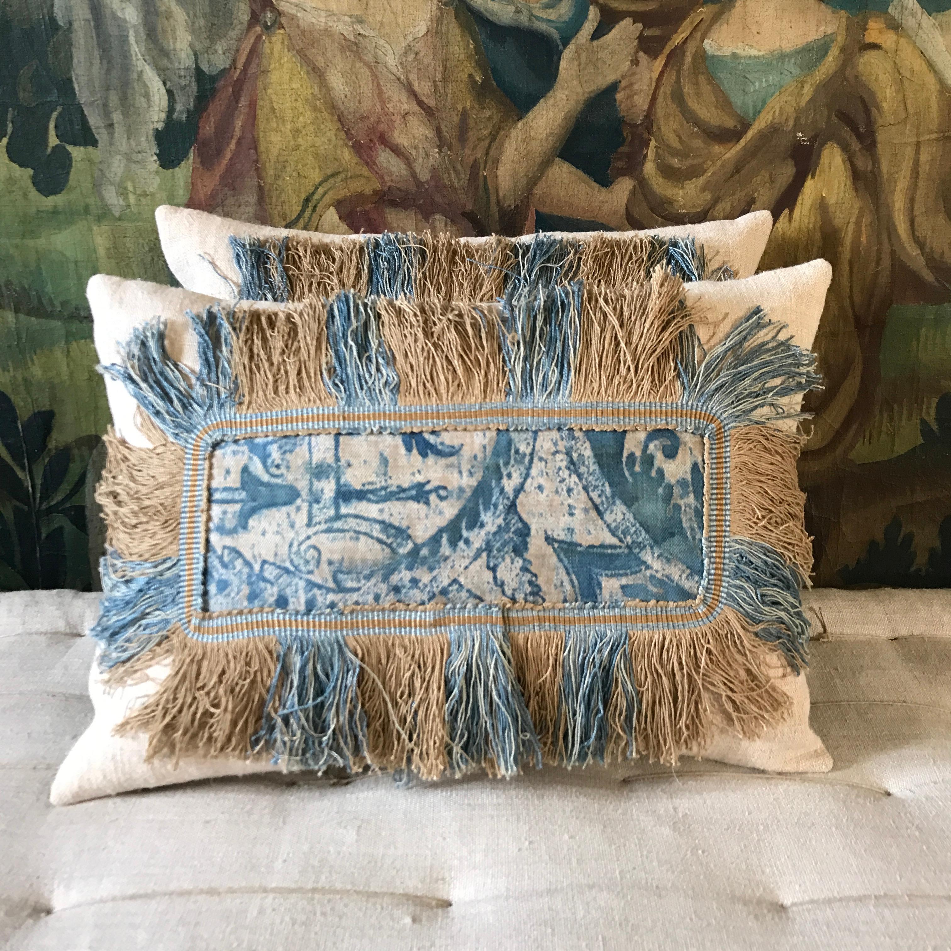 A pair of small cushions made from two fragments of a rare early Fortuny panel circa 1910-20 in a beautiful indigo blue placed on antique linen then trimmed on all edges with a complimentary coloured deep Claremont fringe - with a plain linen to