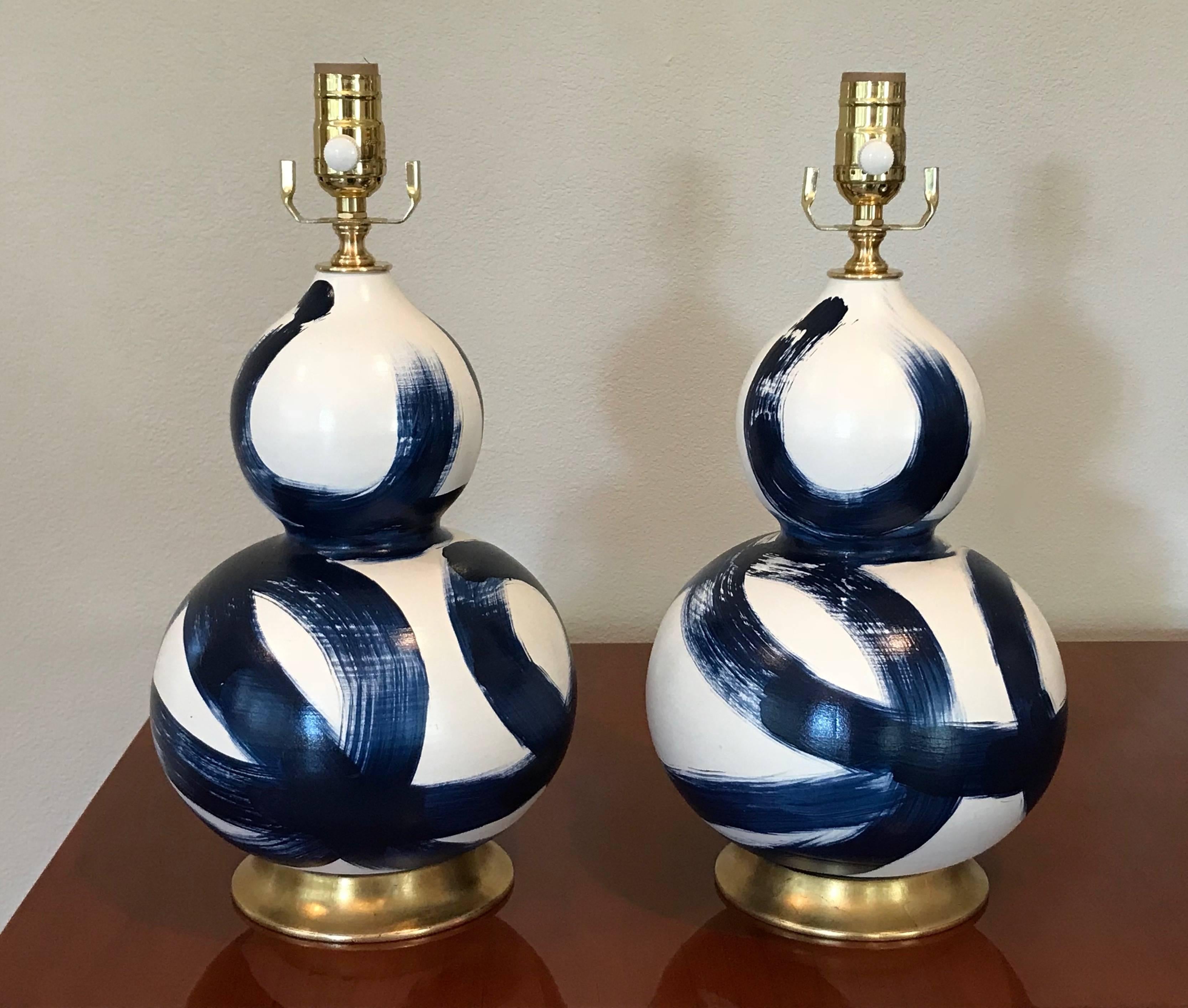 A smaller scale pair of ceramic lamps on turned wood bases in a gilt finish. Lamp bodies are bisque-fired with a matte off-white glaze and dark blue abstract blue brushed design, applied by hand. Newly wired for US with brass fittings and a full