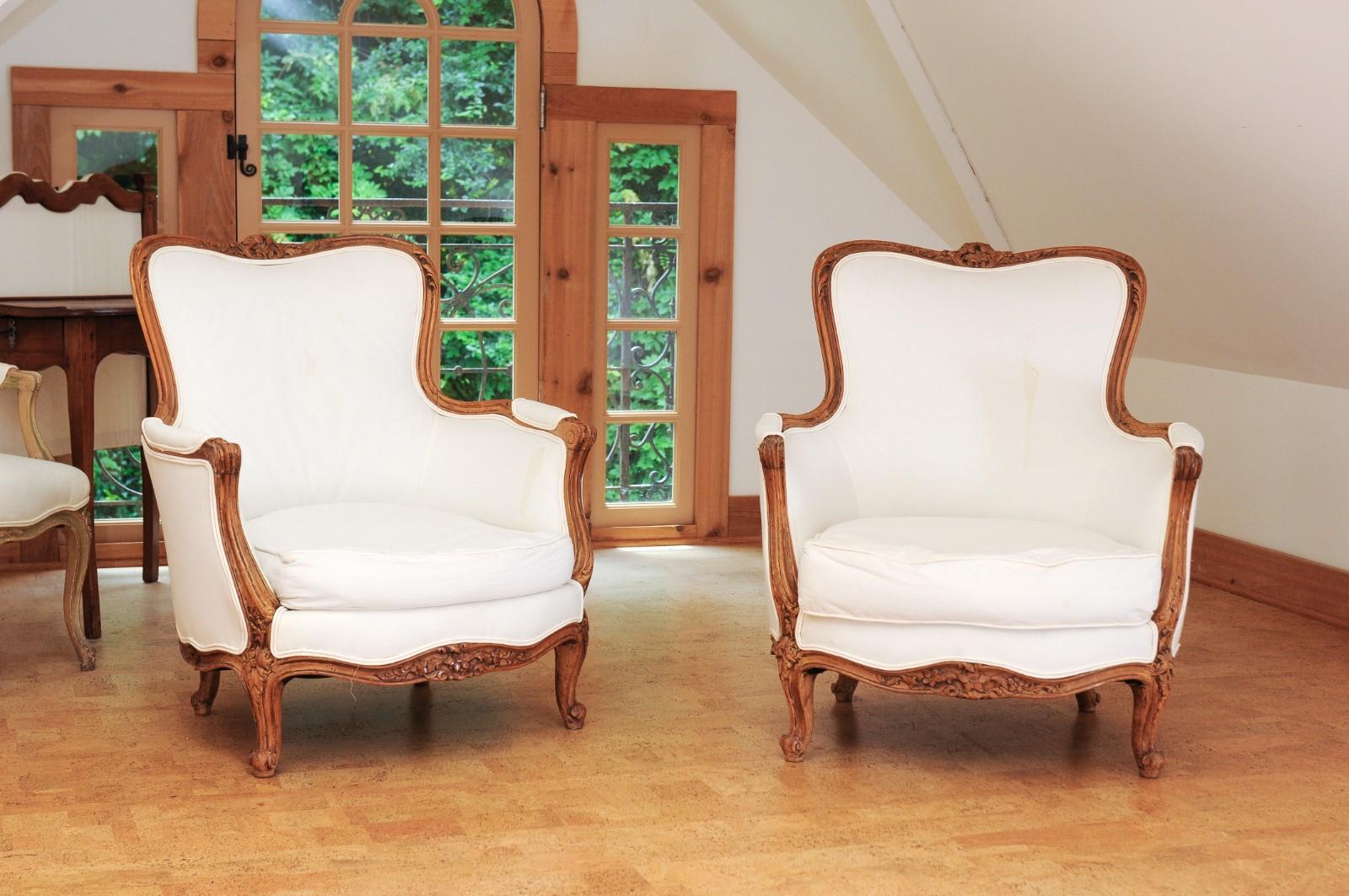 A pair of small French Louis XV style walnut bergères chairs from the 19th century, with upholstery. Created in France during the 19th century, each of this small pair of walnut bergères features a curving back topped with carved foliage. Two