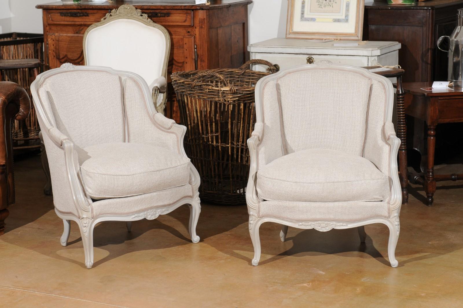 A small pair of French Louis XV style painted wood bergère à oreilles chairs from the 20th century, with floral motifs, cabriole legs and new upholstery. Created in France during the 20th century, each of this pair of petite bergères features the