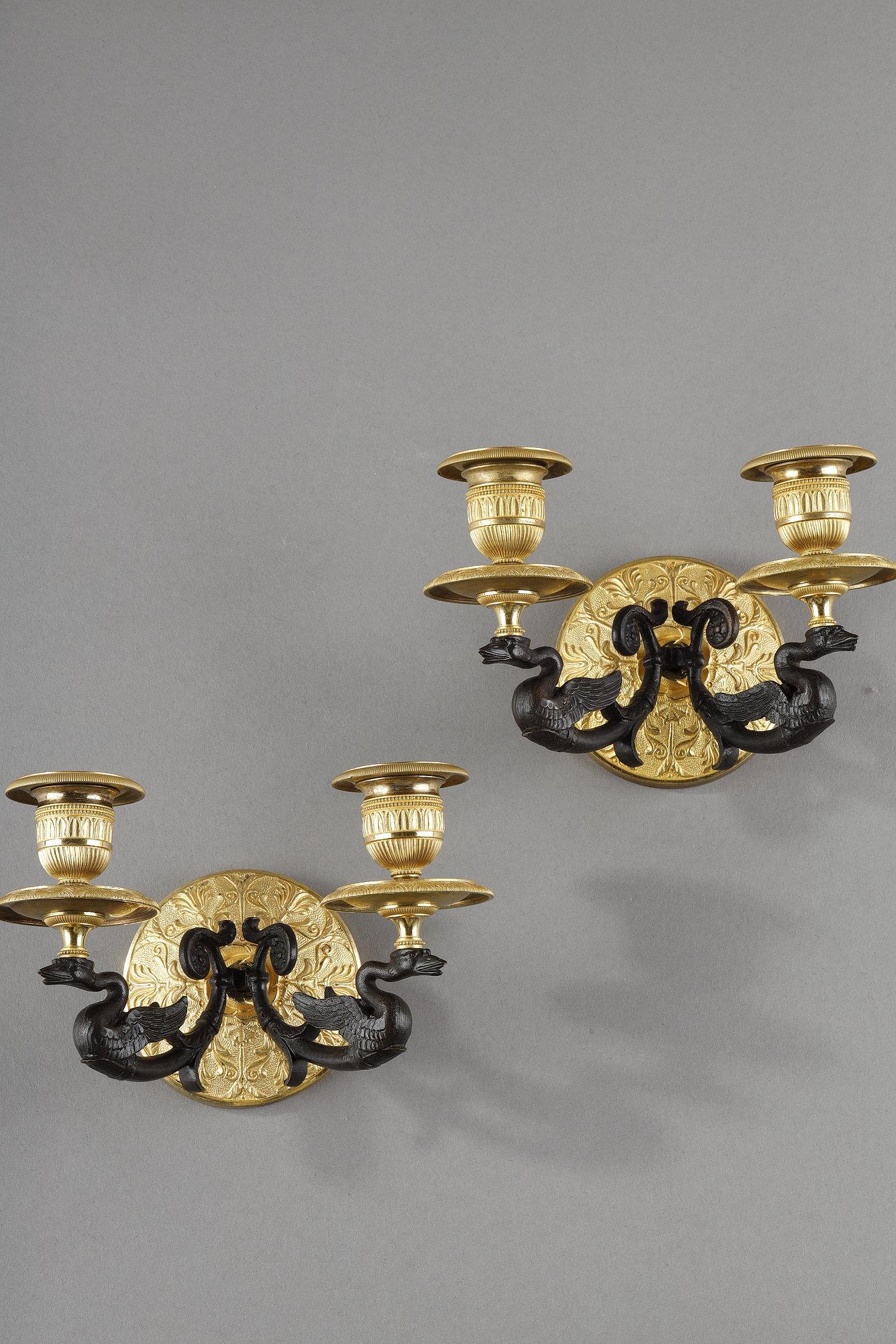 Coppia di appliques in ormolu e patina marrone, con due bracci di luce, a forma di cigno che sostiene lo stoppino sottolineato da un fregio di foglie d'acqua, la base rotonda con palmette.