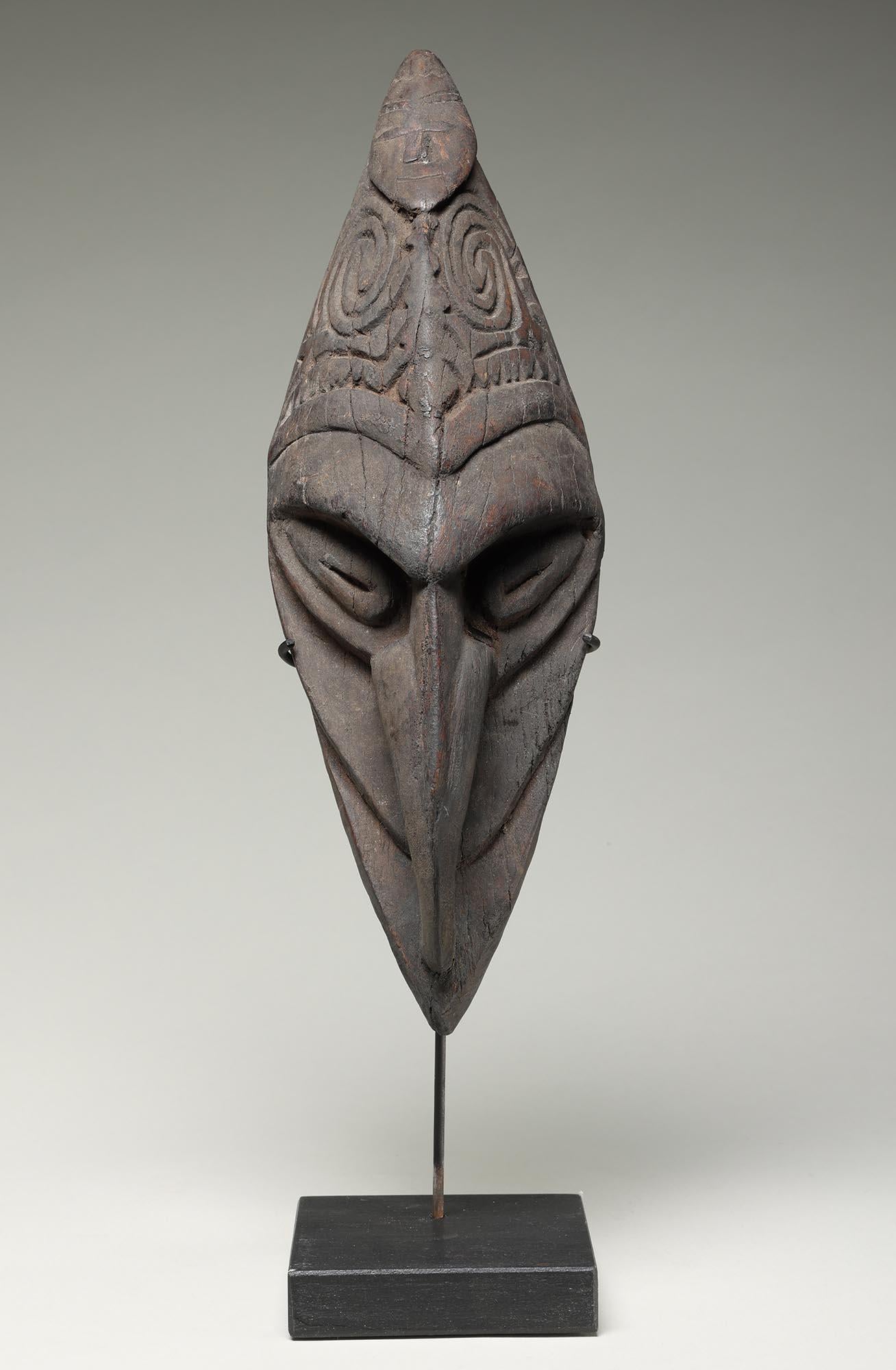 Petit masque d'oiseau en bois foncé sculpté avec un long nez/bec.  Figure plus petite, accroupie, aux bras recroquevillés, sculptée sur le front.  De la région de Sepik River en Papouasie-Nouvelle-Guinée.   Signes d'usure, de saleté incrustée et de