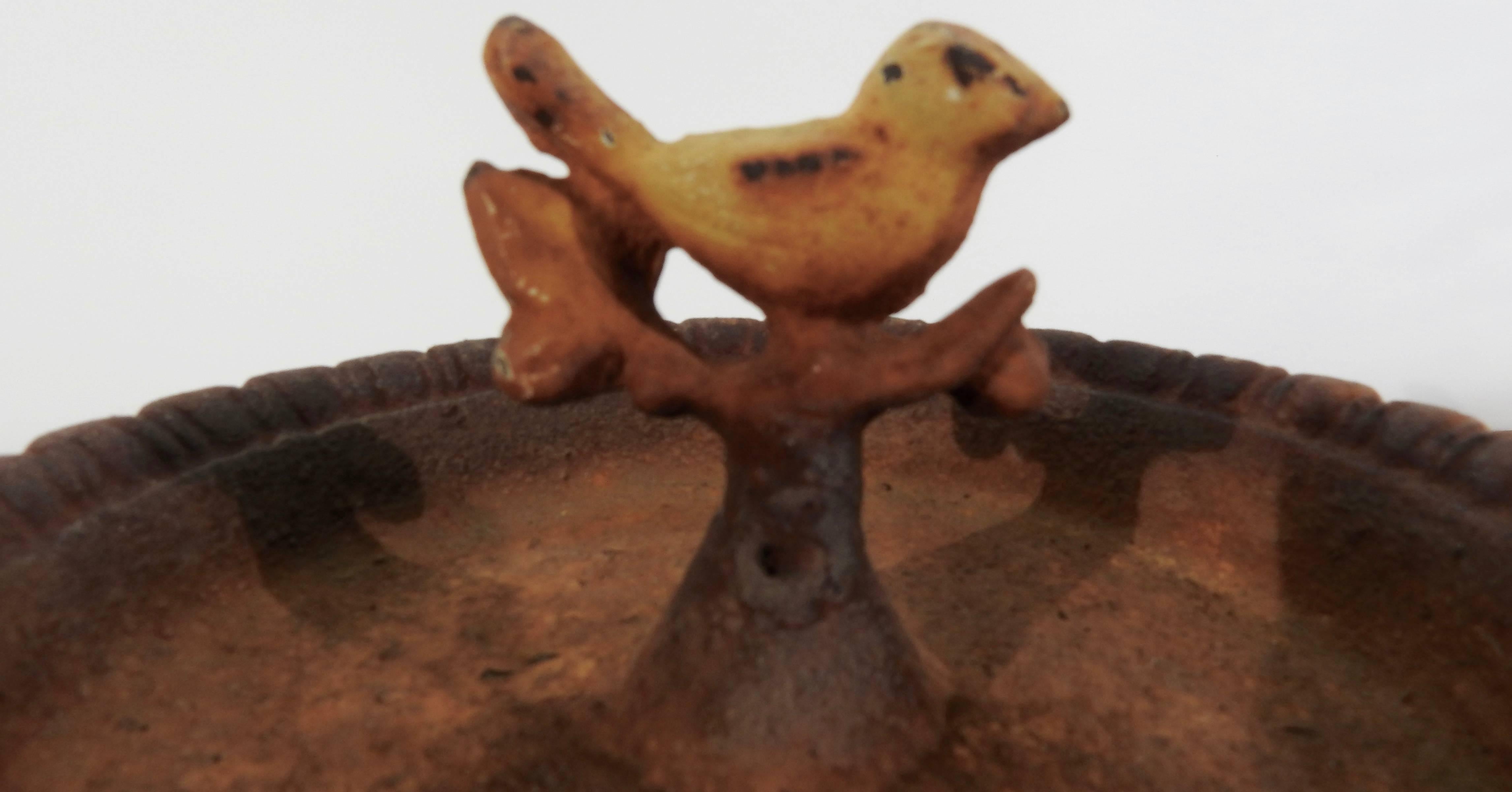 A chipper bird sits atop a branch in the centre of this miniature birdbath. It is made of cast iron that was finished in a white color and one time and now has a rusty, aged patina. This would make a beautiful addition to your surroundings, both