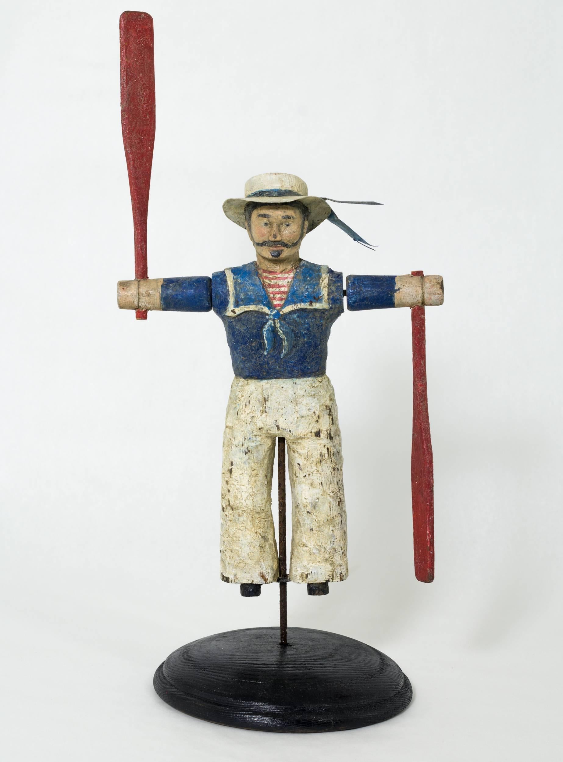Figurilla marinera de pie, tallada en pino que lleva un sombrero blanco con ala de metal,
un jersey azul de marinero con pañuelo, camiseta interior a rayas rojas y blancas, pantalones blancos y zapatos negros. La cara está hábilmente tallada con