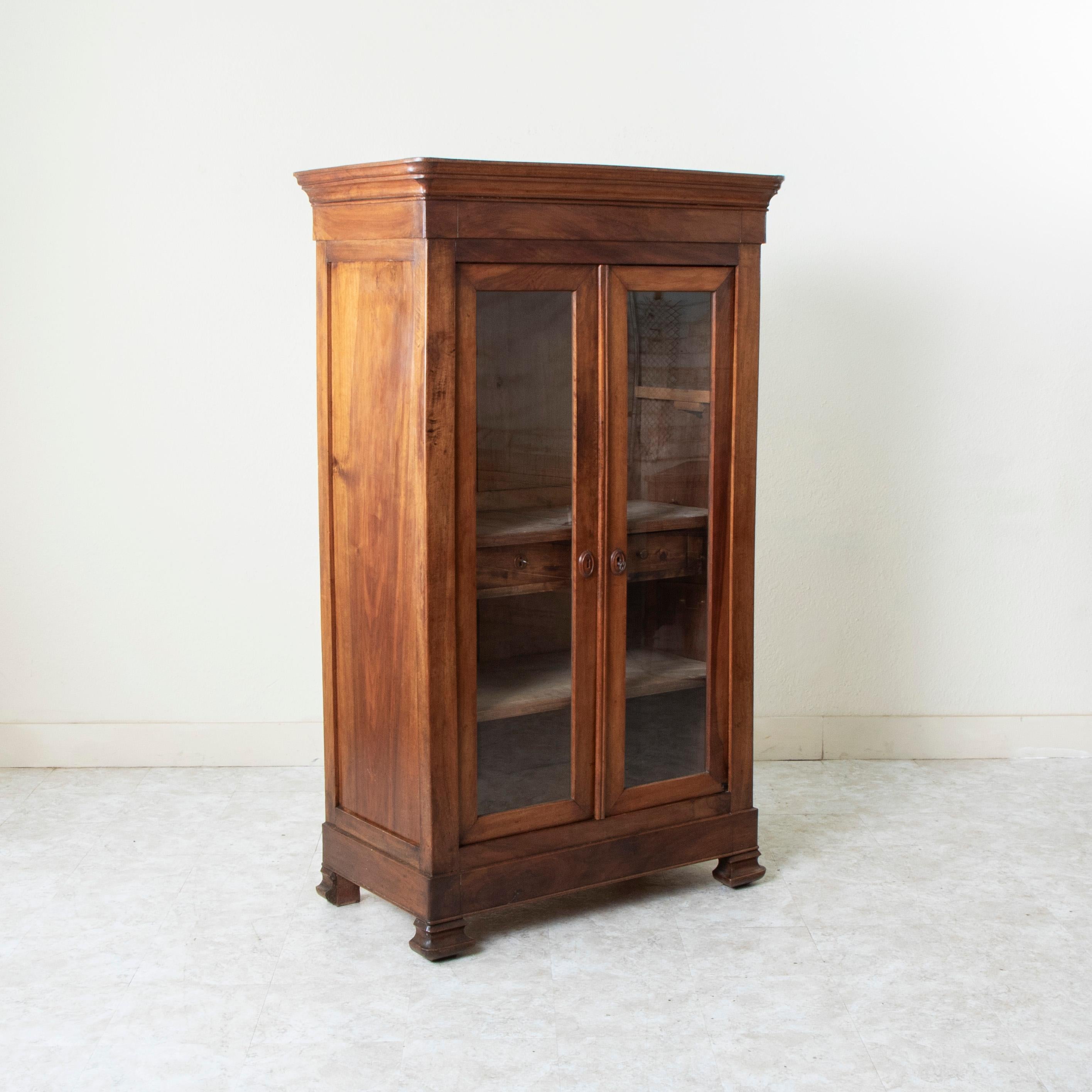 This small scale nineteenth century Louis Philippe period vitrine or bookcase features a facade of solid walnut. Its two glass doors open up to its cabinet with three removable shelves. One of the shelves is fitted with two small drawers of dovetail