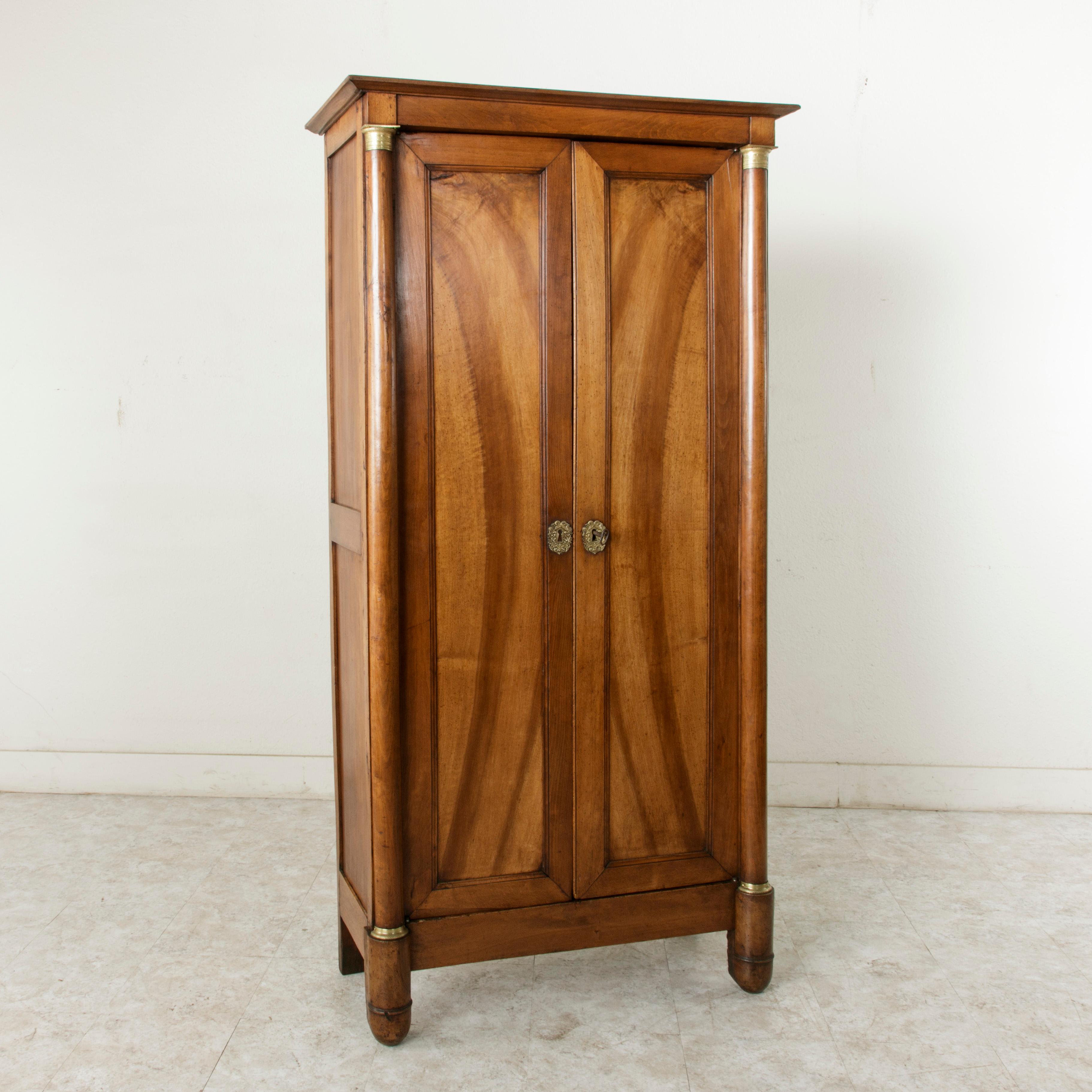 This small scale walnut French Empire period armoire or armoirette features double doors flanked by half columns that are fitted with chased bronzes at the bases and capitals. Bronze escutcheons detailed with an acanthus leaf motif appoint the