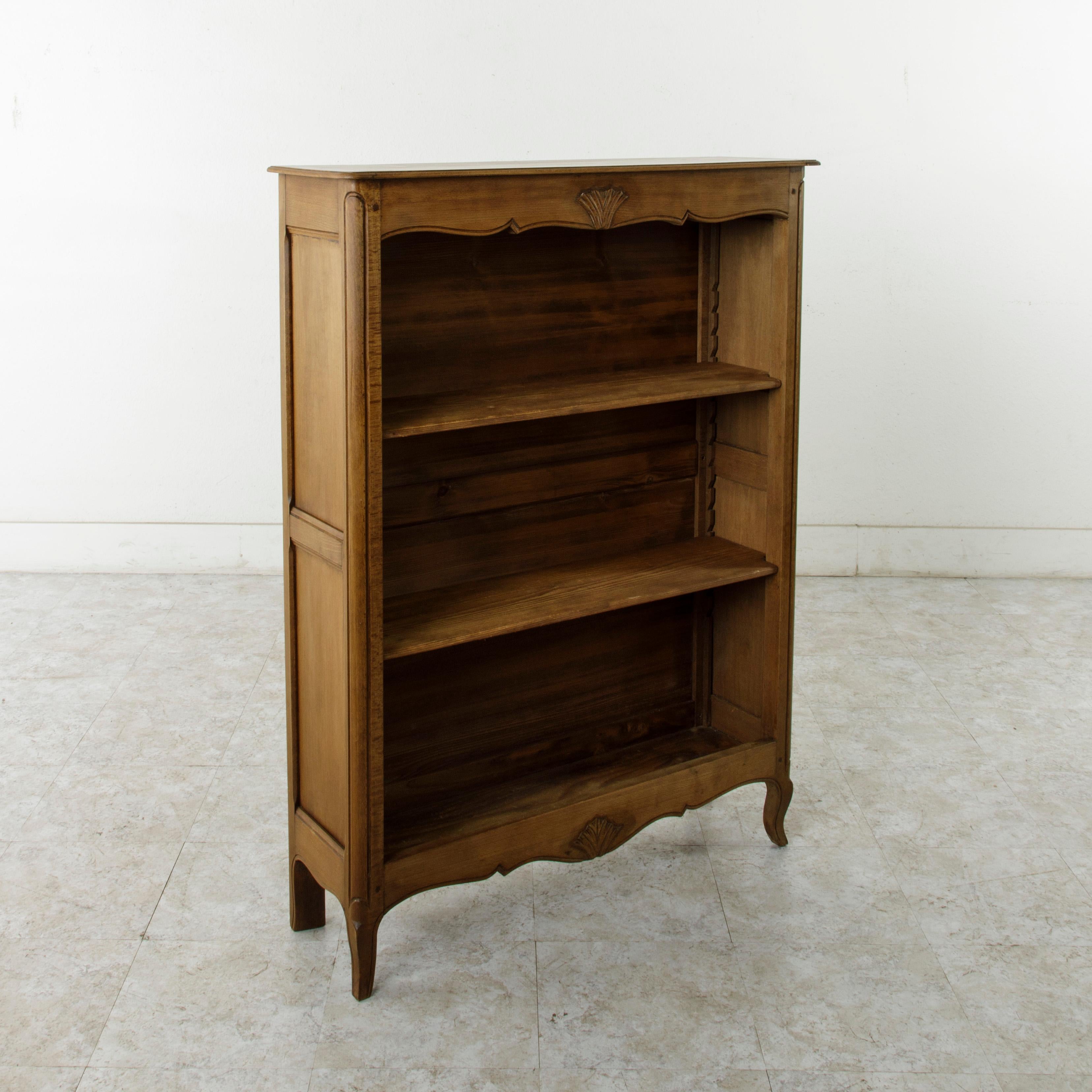 This small scale French Louis XV style bookshelf from the early 20th century is constructed of hand pegged elm with paneled sides. It features a beveled top and fluted corners that rest on cabriolet legs. A central hand carved leaf adorns the upper