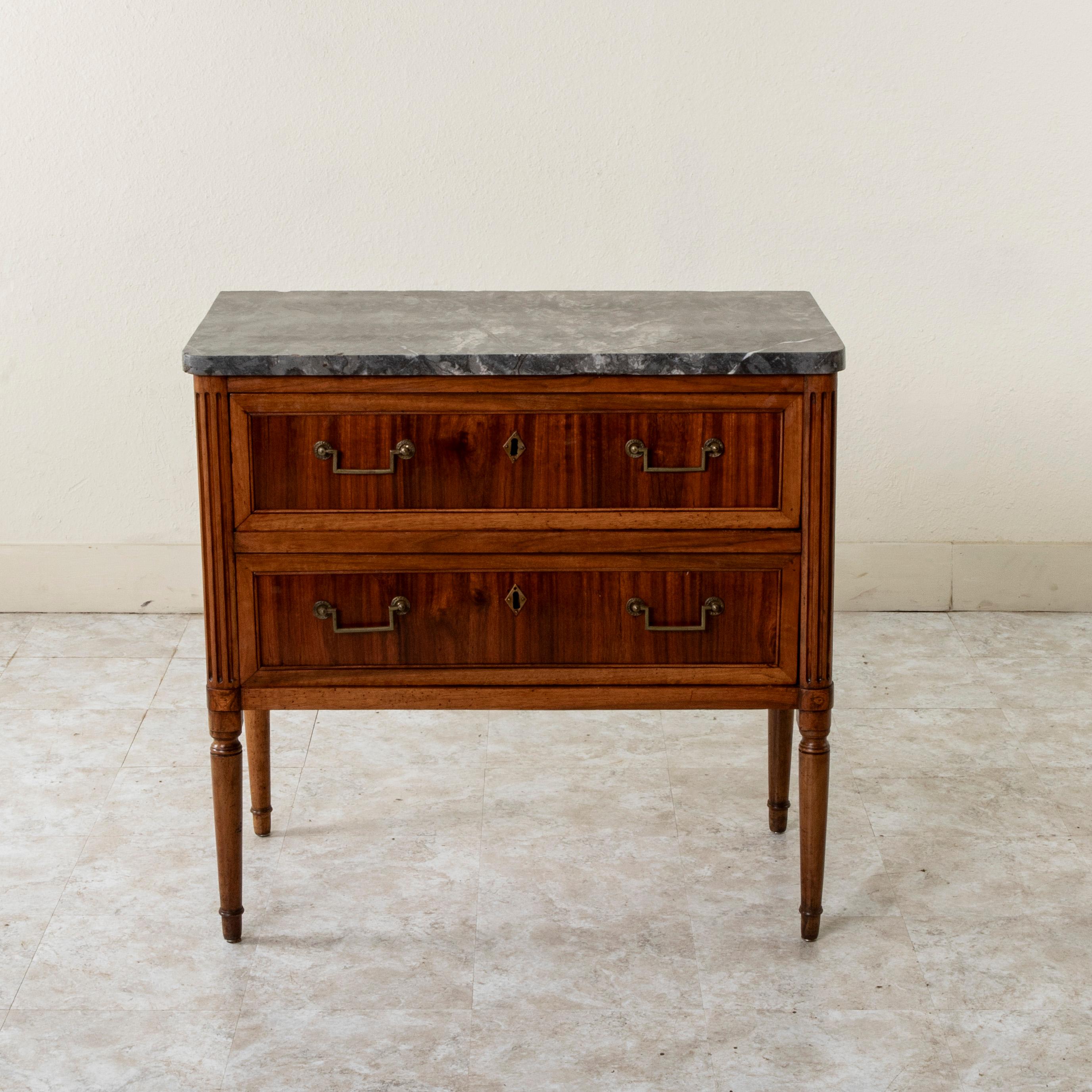 Measuring only 34.5 inches wide, this small scale late eighteenth century French Louis XVI period walnut commode or chest of drawers features a grey marble top and fluted rounded corners. Its two drawers of dovetail construction are detailed with