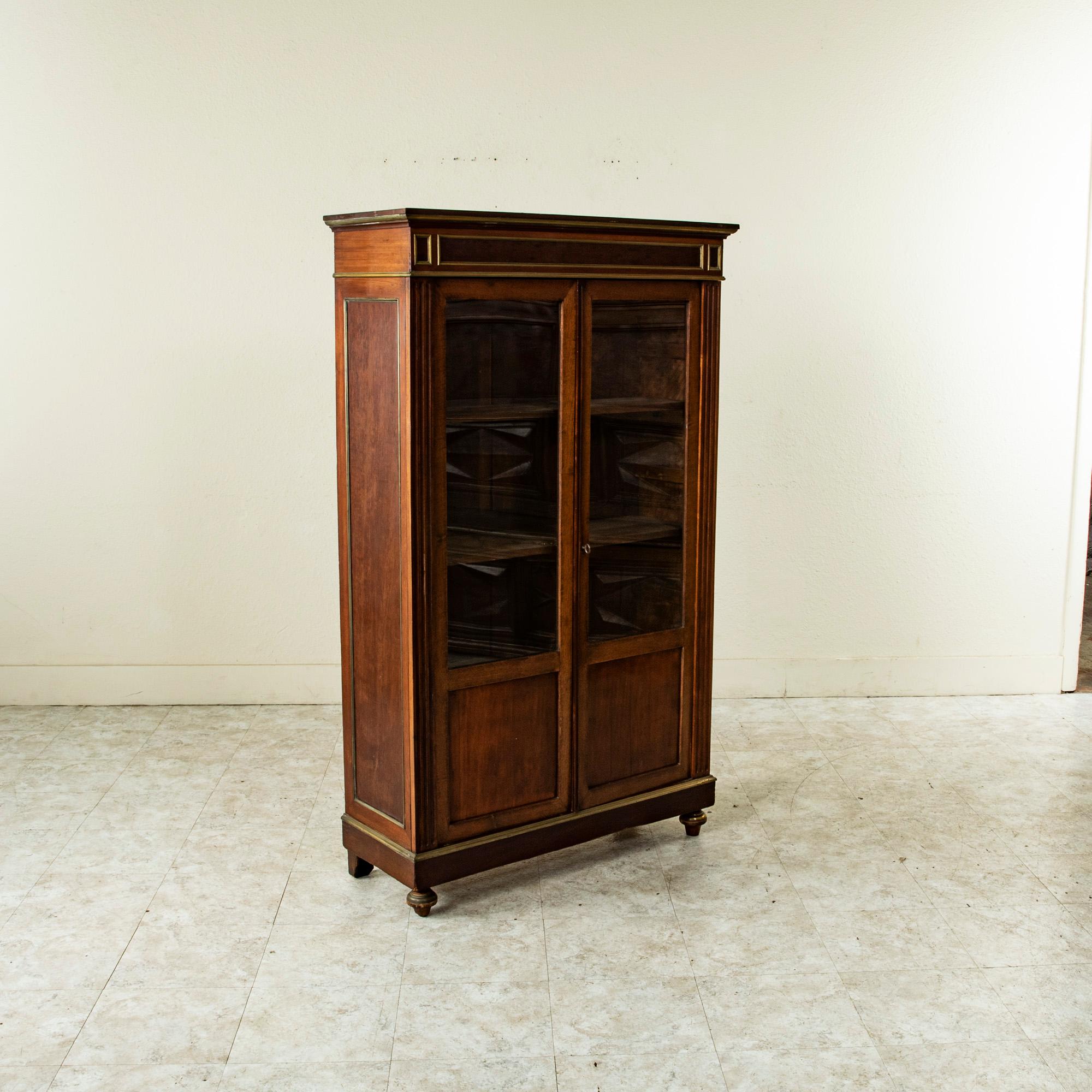 This small scale late 19th century French Louis XVI style mahogany bibliotheque or bookcase is finished with bronze banding. Its corners feature fluting and the side panels are also inset with bronze banding. Its two doors open to reveal an interior
