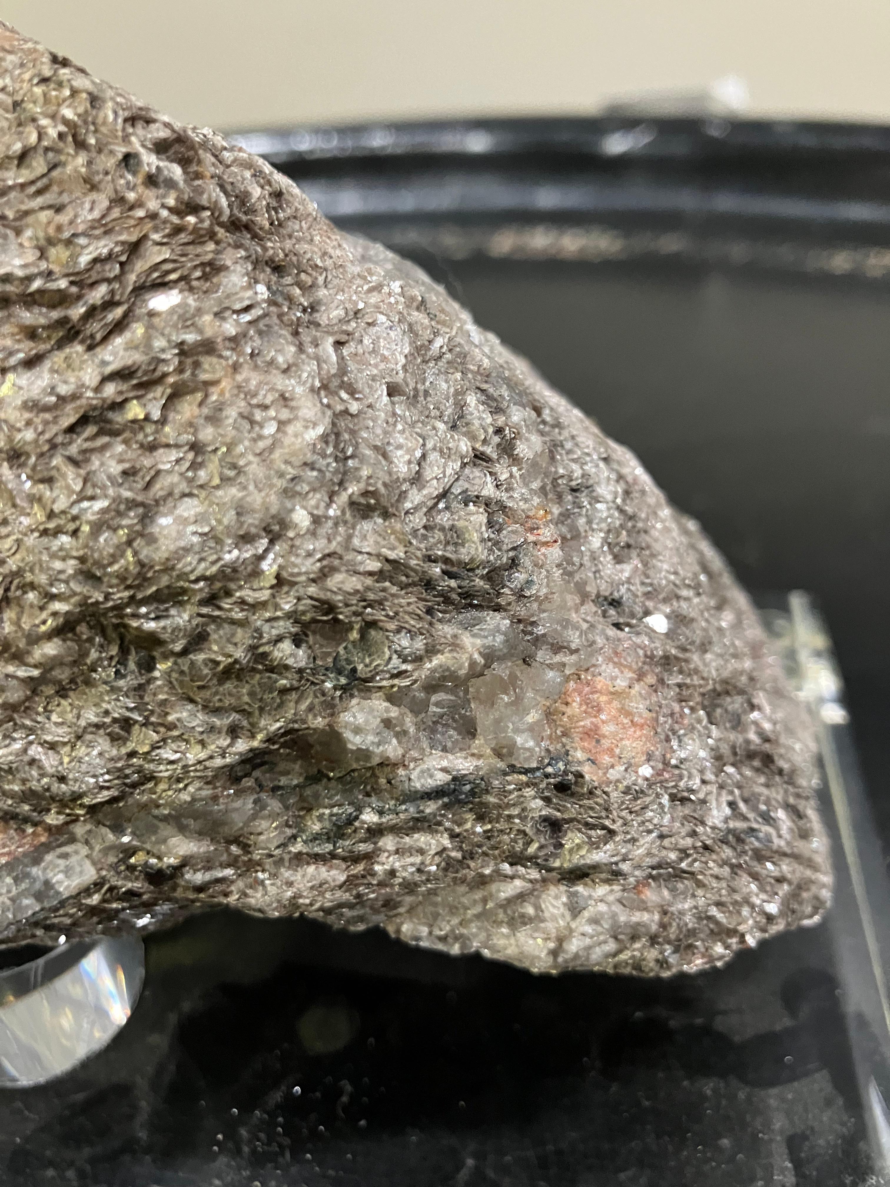 A compact, naturally textured selenite specimen mounted on a clear acrylic block for presentation. The stone features layered, fibrous formations with a subtle shimmer, ranging in earthy tones from silvery gray to mineral brown with hints of pale