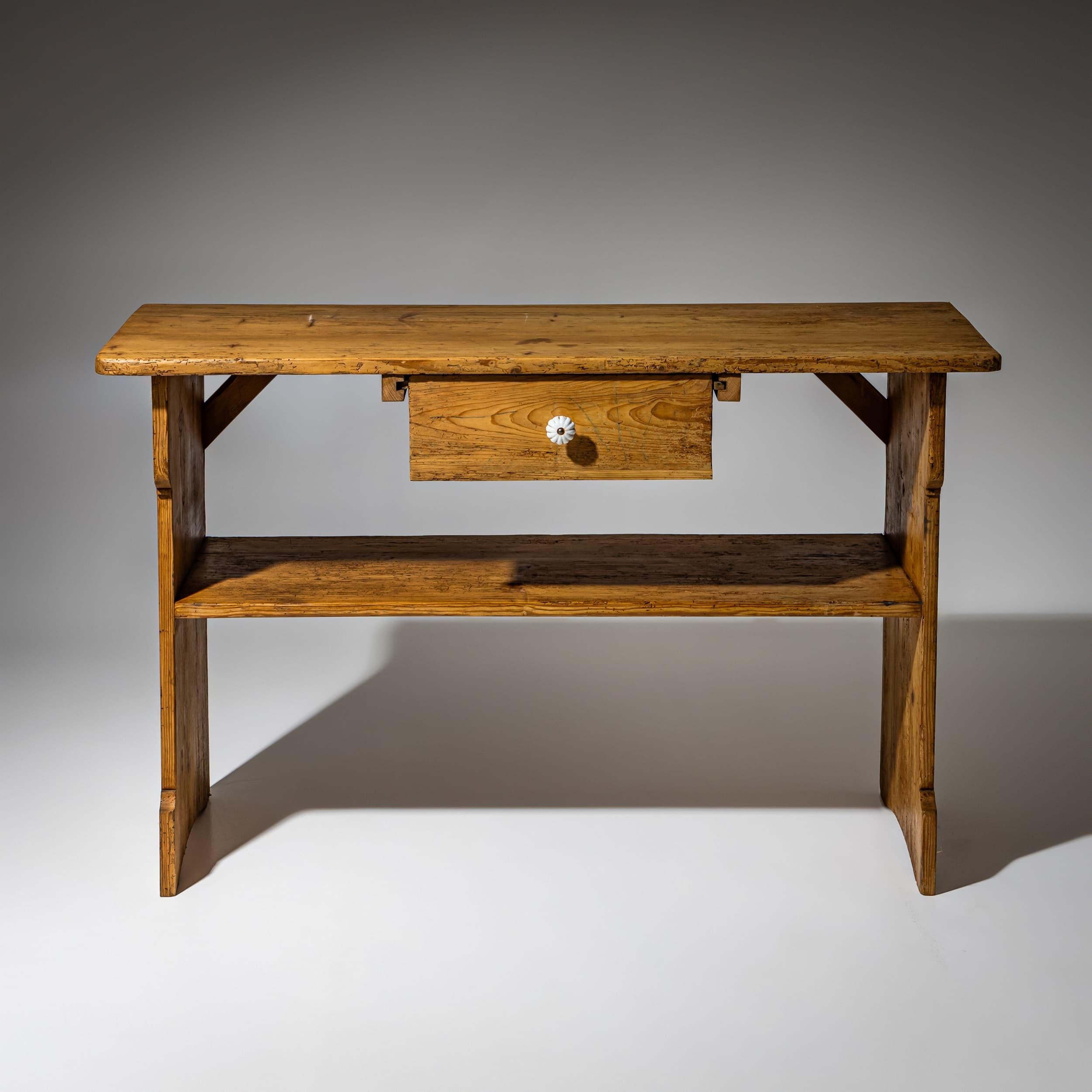 Table d'appoint en sapin, de facture simple, avec plateau droit, panneaux latéraux à tenons traversants et profil inférieur façonné. Tiroir en position centrale sous le tablier, équipé d'un bouton en céramique. Une étagère inférieure encastrée