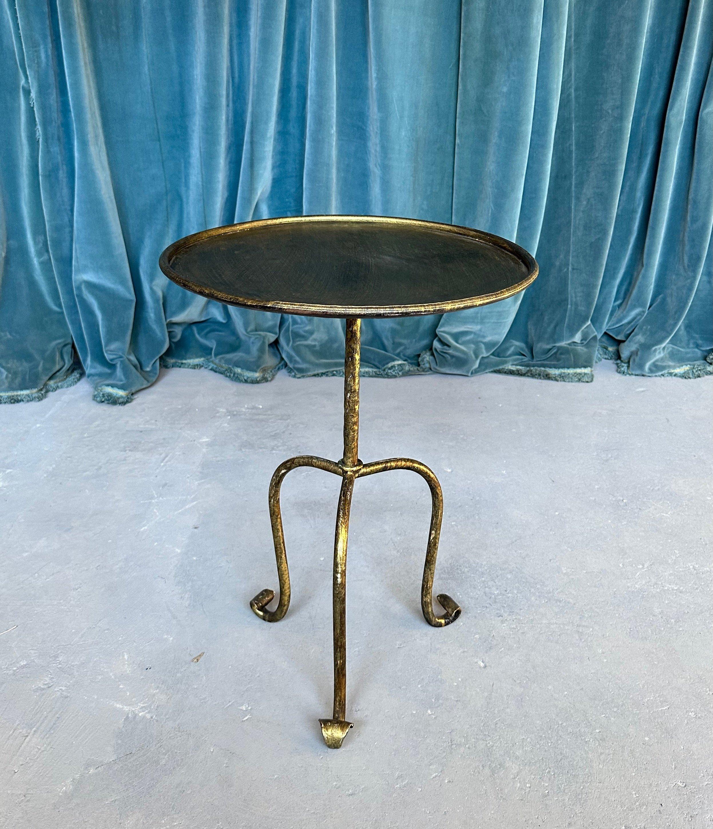 This unique small scaled side table was recently made by European artisans. It features a hand applied gold finish with dark undertones and is mounted on a fanciful elevated tripod base ending in upwardly scrolled feet. The size of this table is