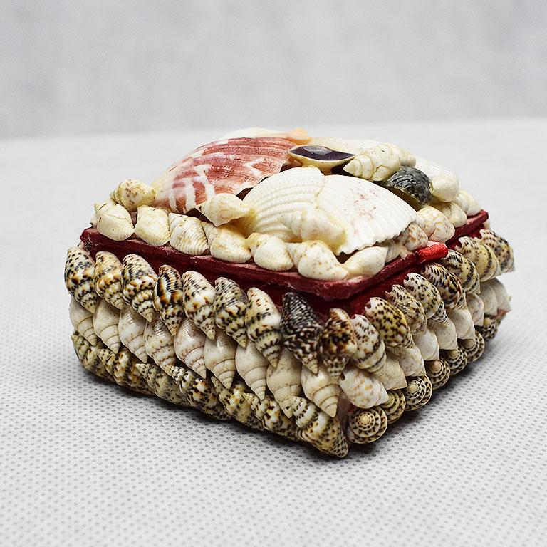 Small square shell encrusted trinket box. Covered in a variety of shells, this box opens to reveal a red-lined fabric interior. This would be the perfect piece to display on a coffee table or vanity to hide jewelry or other trinkets or jewelry.
