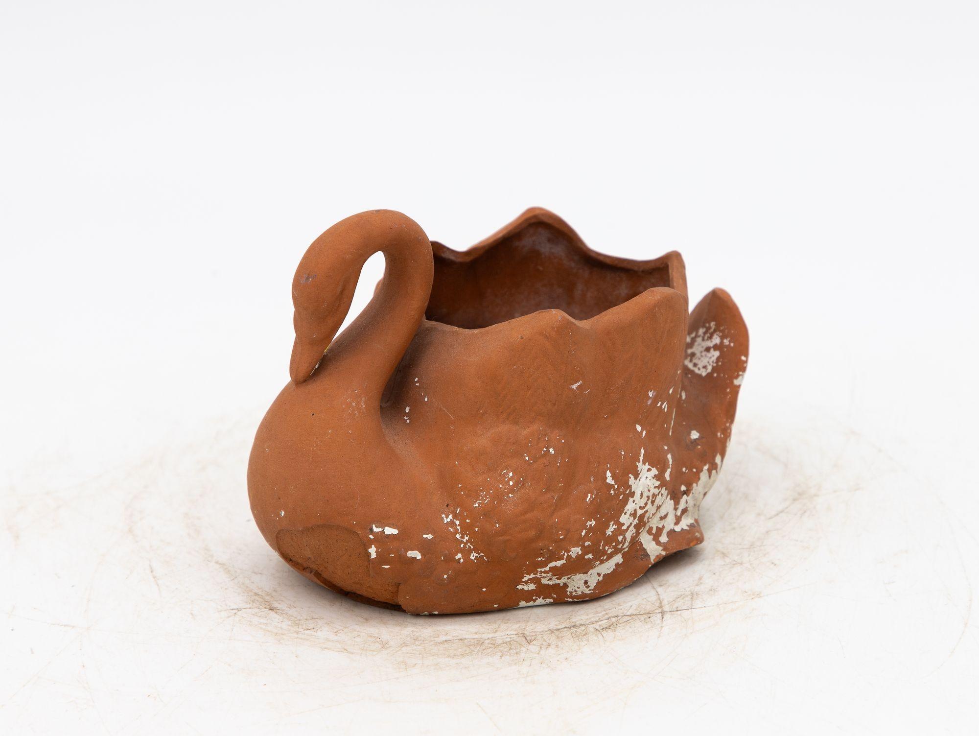 This small tabletop terracotta planter, shaped like a swan with a gracefully curved neck, dates back to the late 20th century. It features remnants of white paint and some areas of terracotta loss, adding to its vintage charm. This unique piece