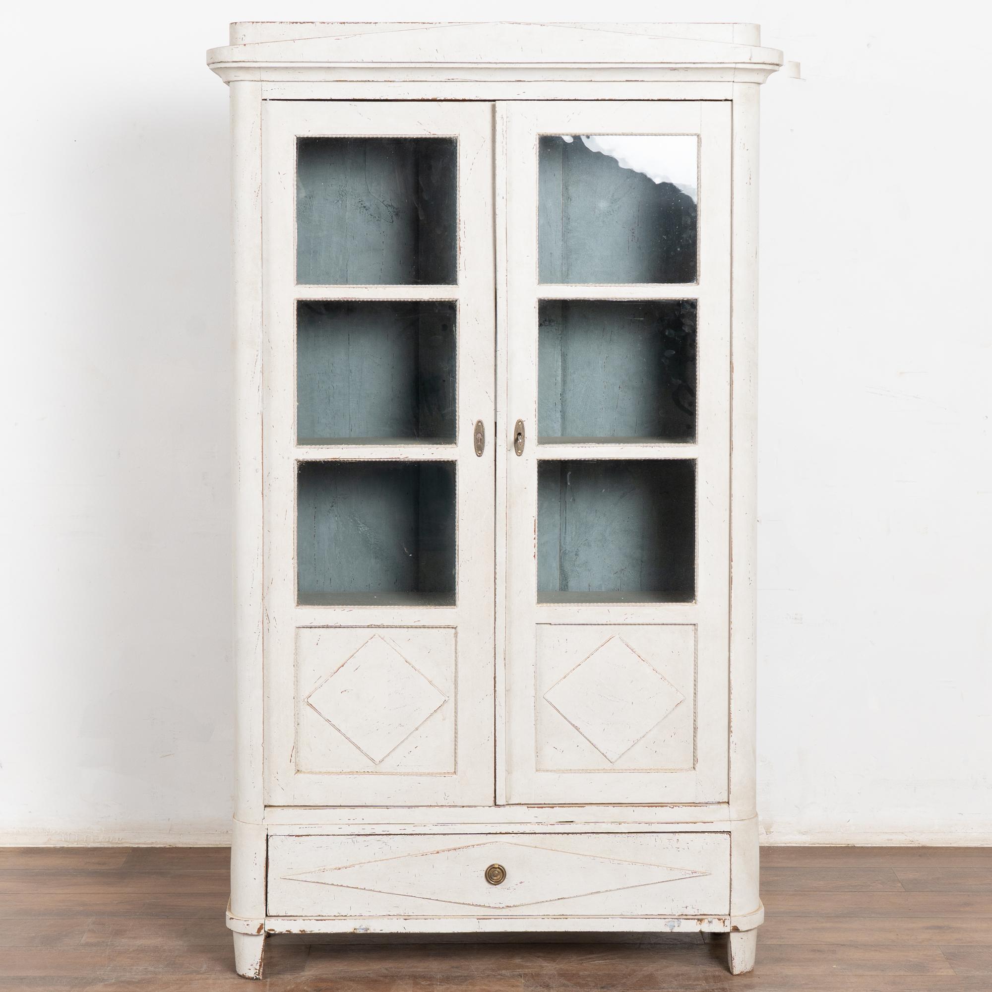 Small Swedish Gustavian pine bookcase featuring a professionally applied layered antique white painted finish with pale gray undertones. Light distressing reveals natural pine wood beneath and fits the age and grace of this cabinet.
The pane glass
