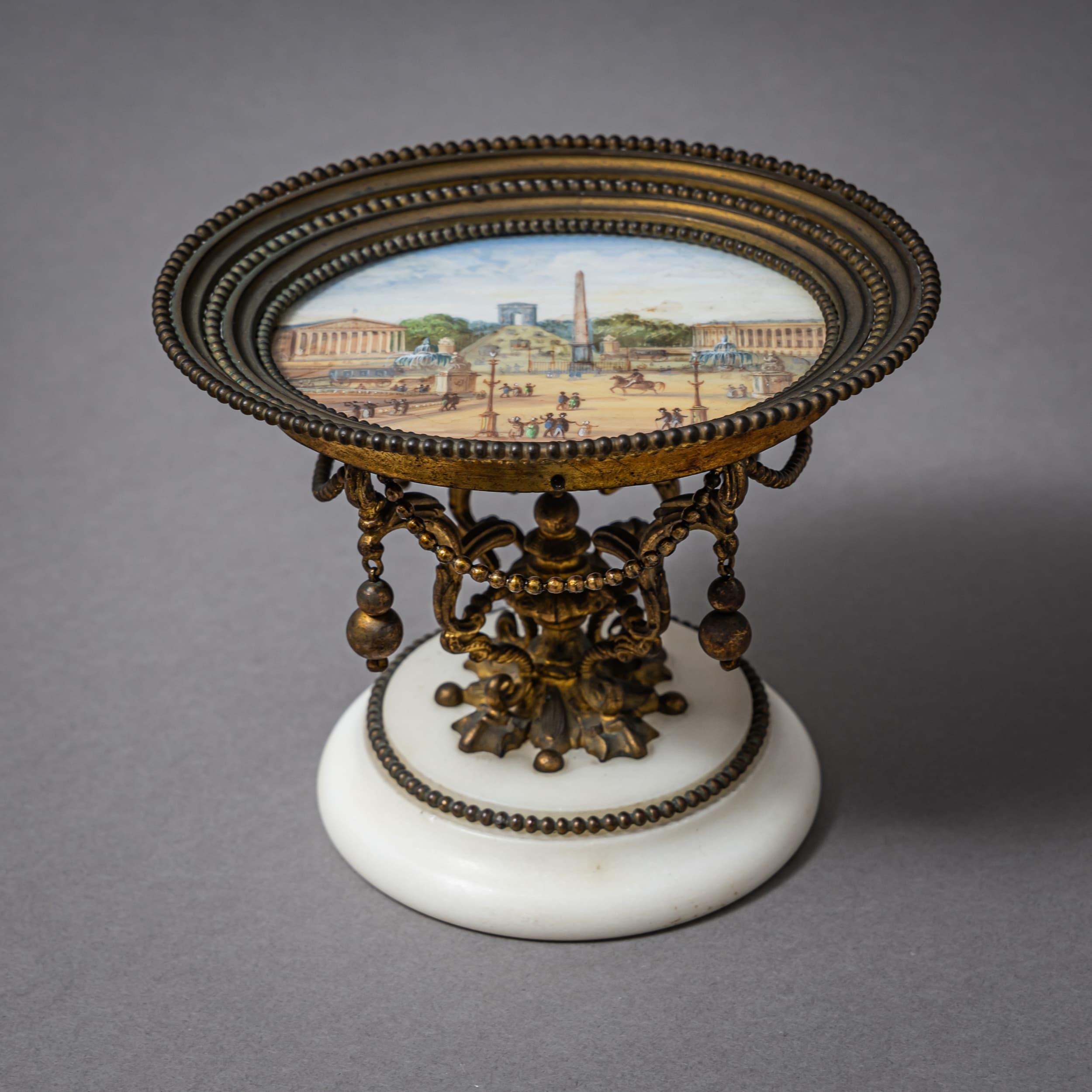 Elegante Tazza mit einem ovalen Podest und einer Porzellanplakette, die eine mehrfarbige Stadtansicht von Paris zeigt. Die Szene zeigt die Place de la Concorde mit dem Arc de Triomphe in der Ferne, dem Palais Bourbon zur Linken und dem Hôtel de