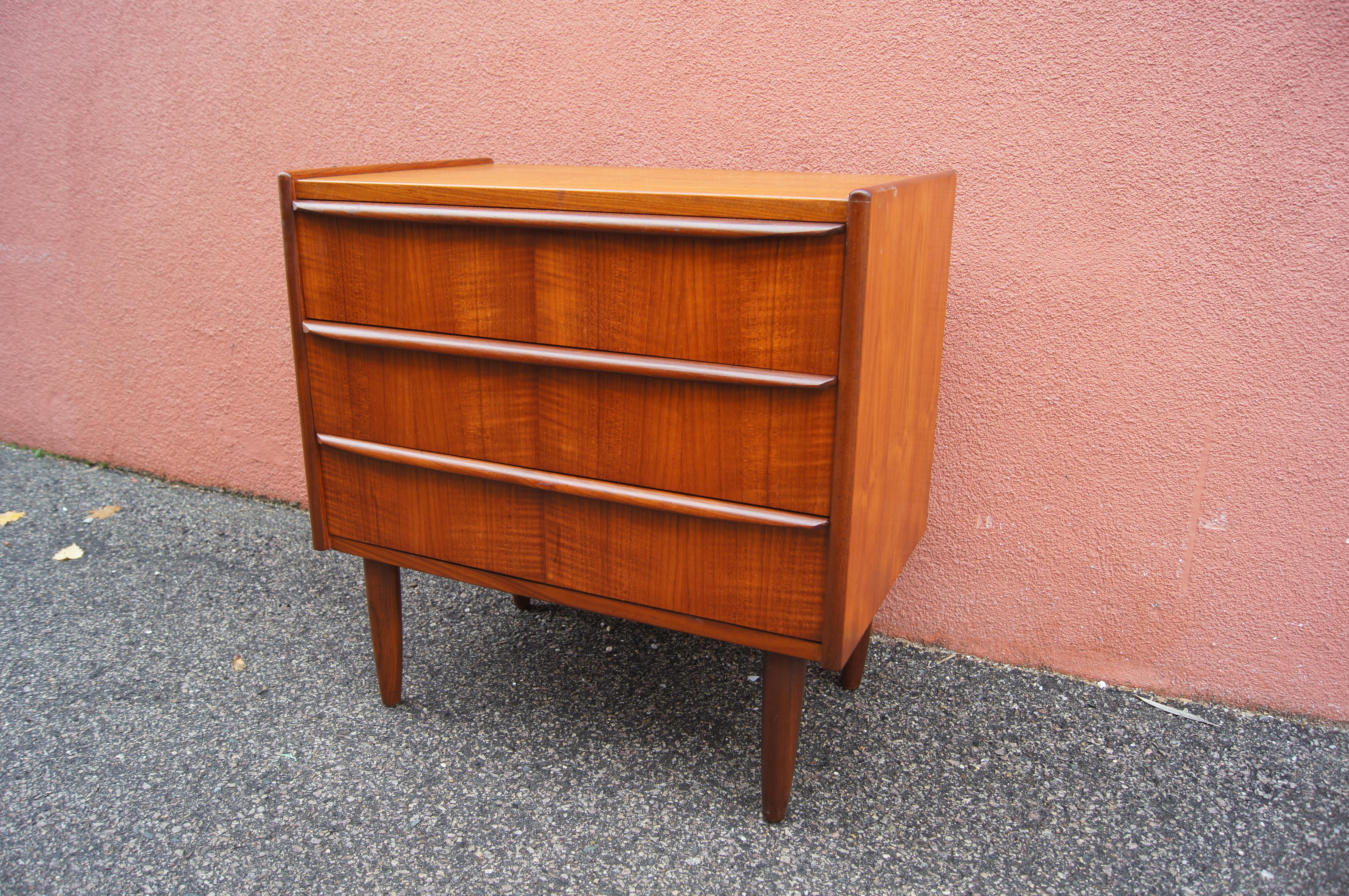 This small Danish modern chest or dresser comprises a case of beautifully grained teak with a raised lip at either side and slightly tapered legs. The three drawers feature integrated linear pulls.