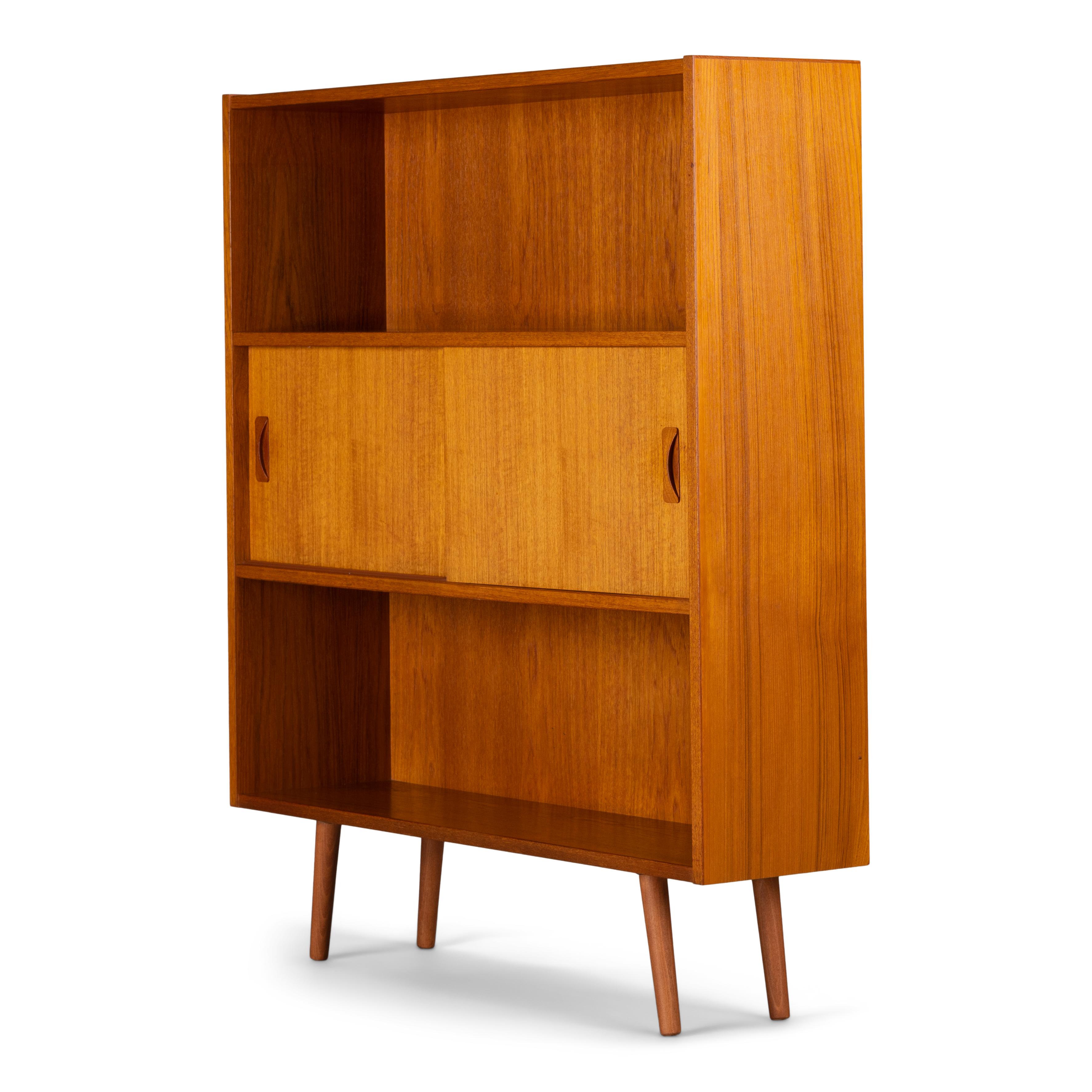 Danish midcentury small bookcase in beautiful teak veneer by Clausen & Søn. This chest from the mid-1970s comes with 2 book shelves and a section with two sliding doors in the middle of the book case. This book case is still original and in a very