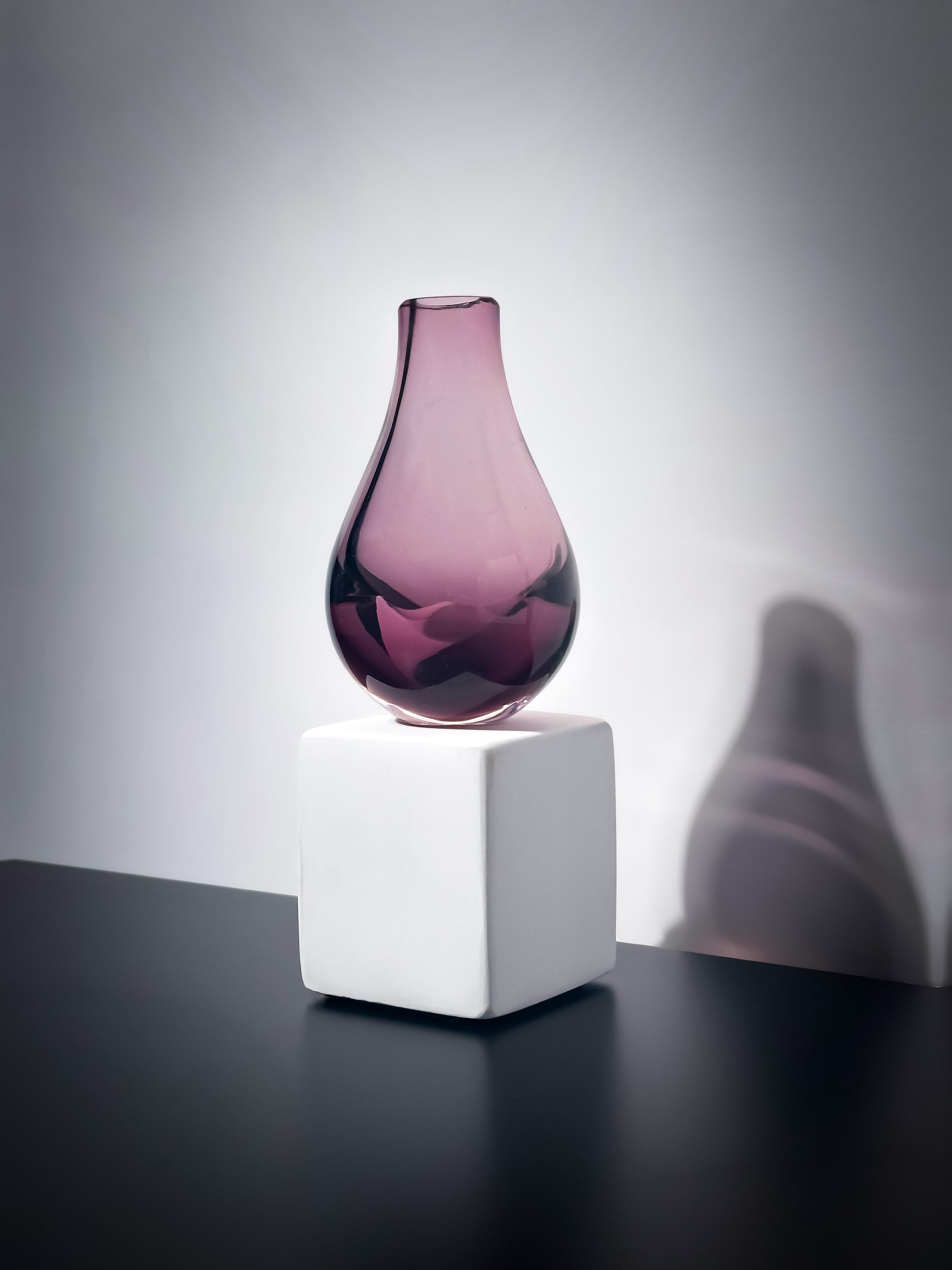 A small art glass bud vase by the Scottish glassmakers Caithness, hand crafted in the 1960s. 

This teardrop-shaped vase, featuring an amethyst-toned body encased in a delicate layer of clear glass, is attributed to Domhnall O'Broin—an influential