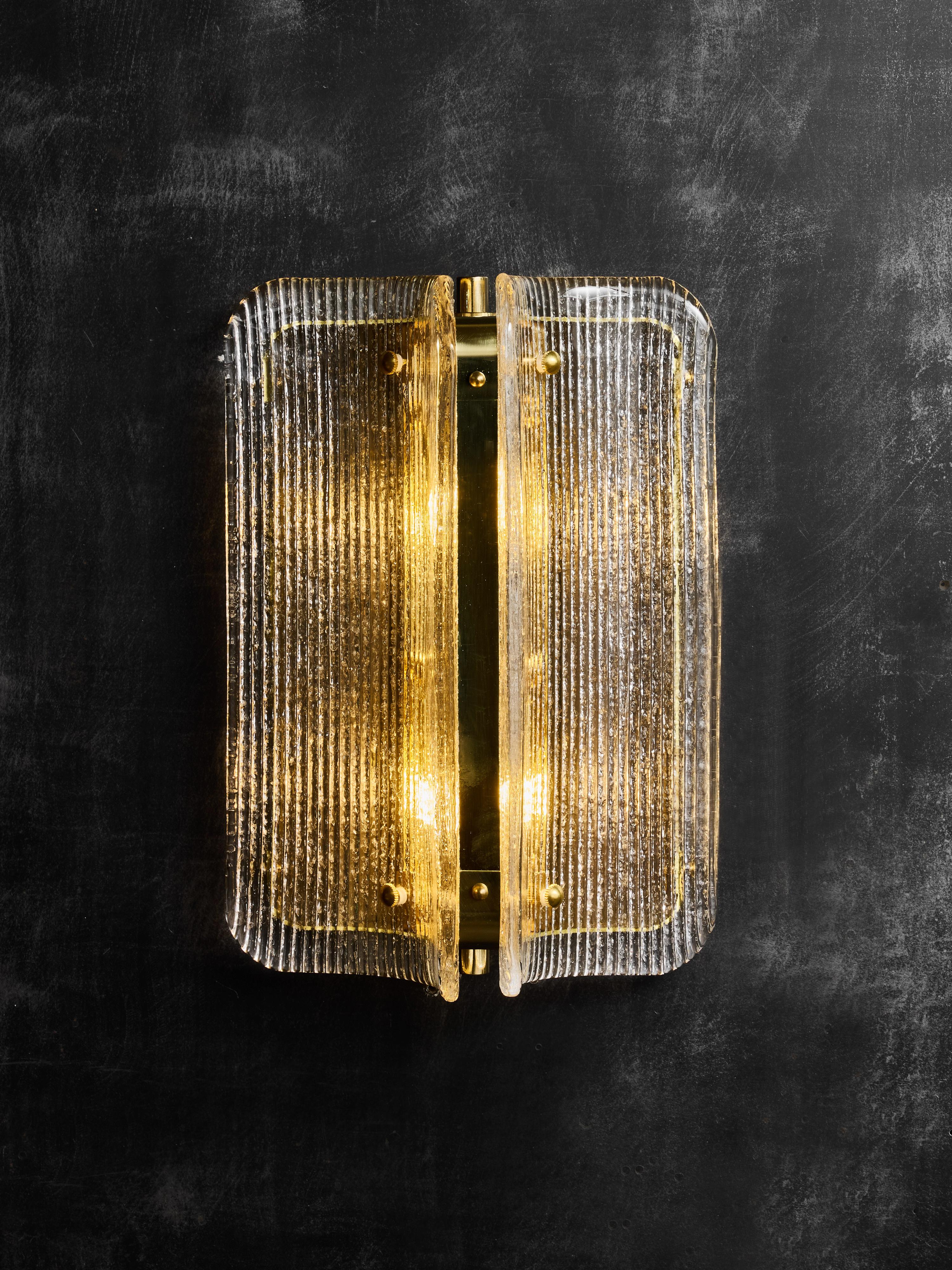 Un sofisticado aplique de pared moderno, con un conjunto escultural de paneles de cristal texturizado montados alrededor de una estructura central de latón. La lámpara se compone de dos elementos de cristal curvados verticalmente, cada uno con una