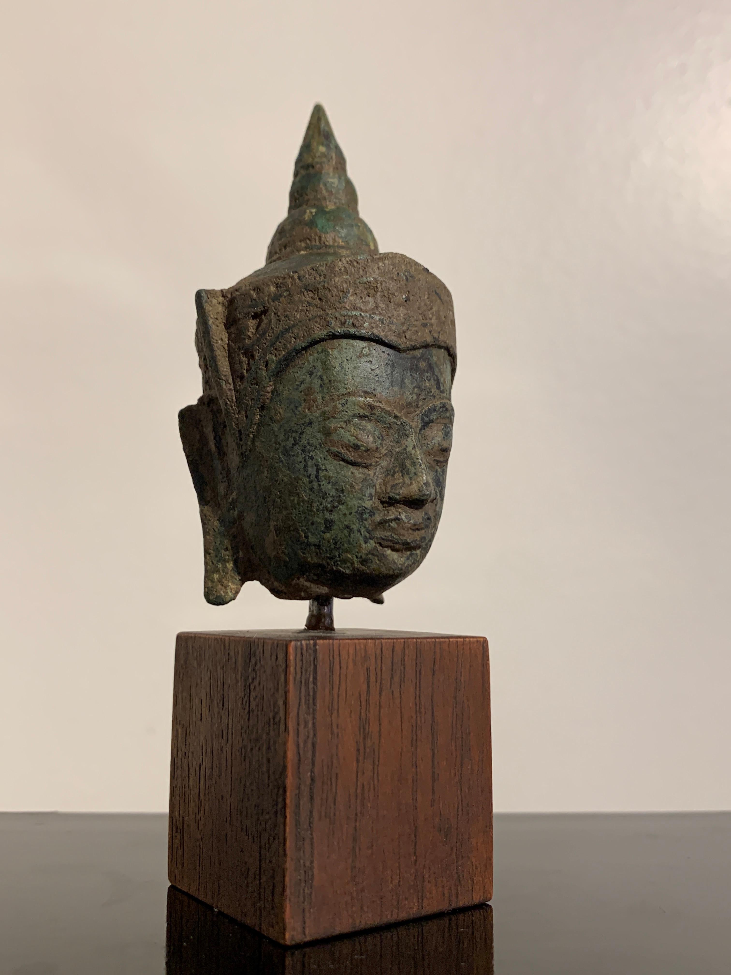 A small cast bronze crowned head of the Buddha with heavy patina, Ayutthaya Kingdom, 17th century, Thailand. 

The full face of the Buddha features heavily lidded downcast eyes, a wide nose, and full lips turned up in a slight smile. Long pendant