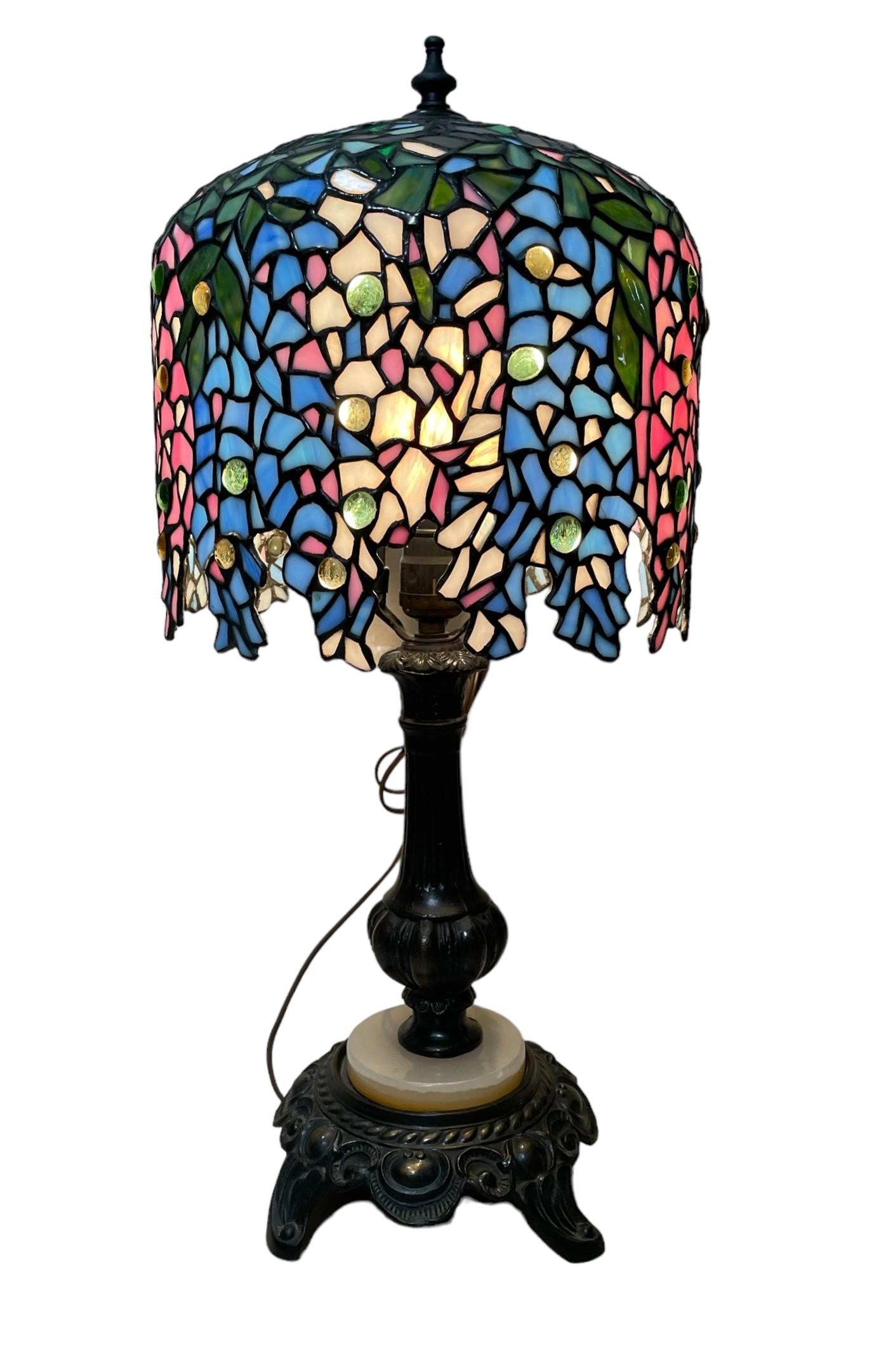 Se trata de una lámpara de mesa de estilo Tiffany. Representa una base de lámpara en forma de balaustre estriado de metal, decorada con rocallas repujadas y pies de volutas en la parte inferior. Una placa redonda de ónice color perla adorna también