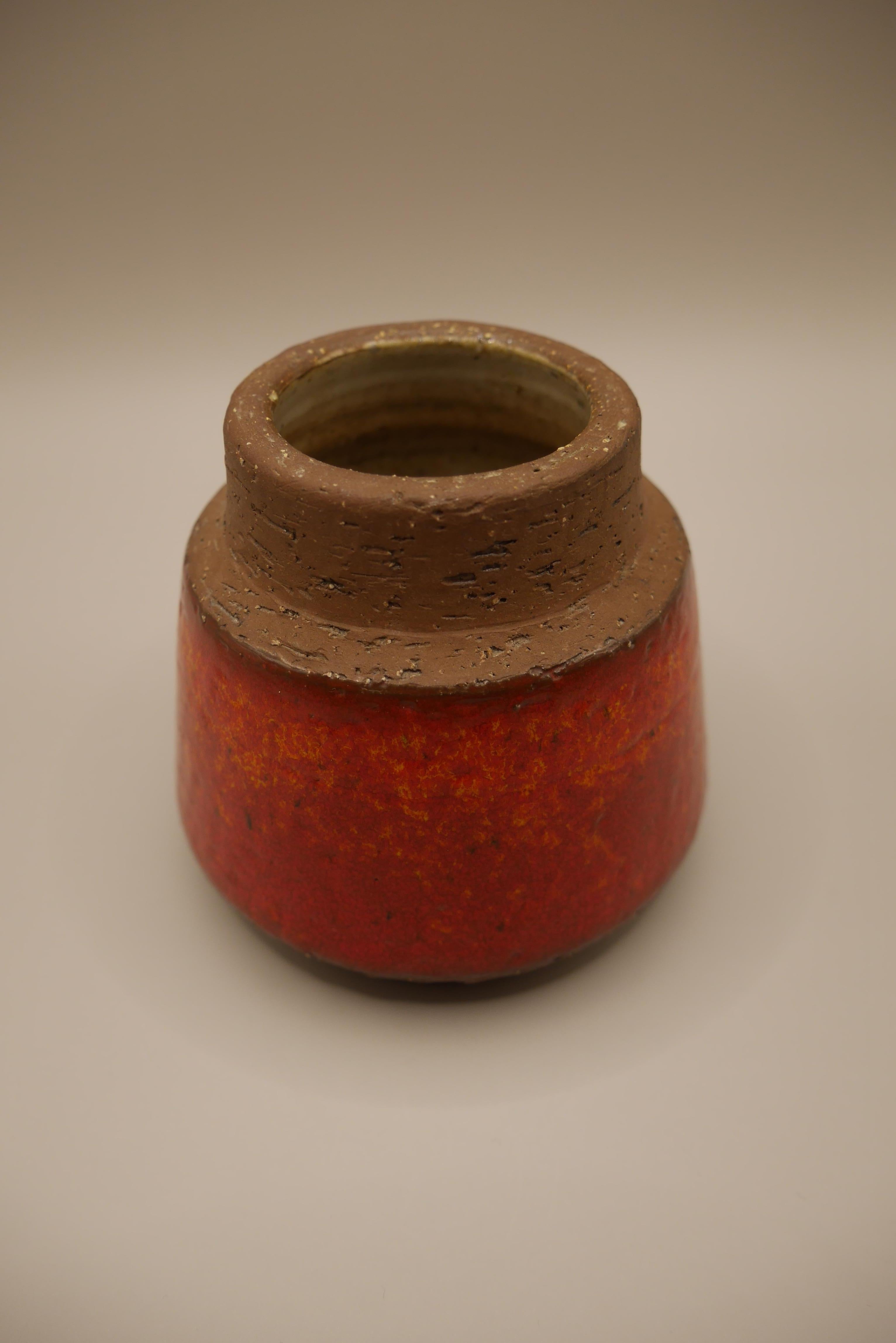 A small vase or pot by Micheal Andersen for Bornholm ceramics in Denmark. Heavy, rough clay with a red/orange glaze around the body of the piece. This vase is part of a series in the same style which featured many variations of the same form with