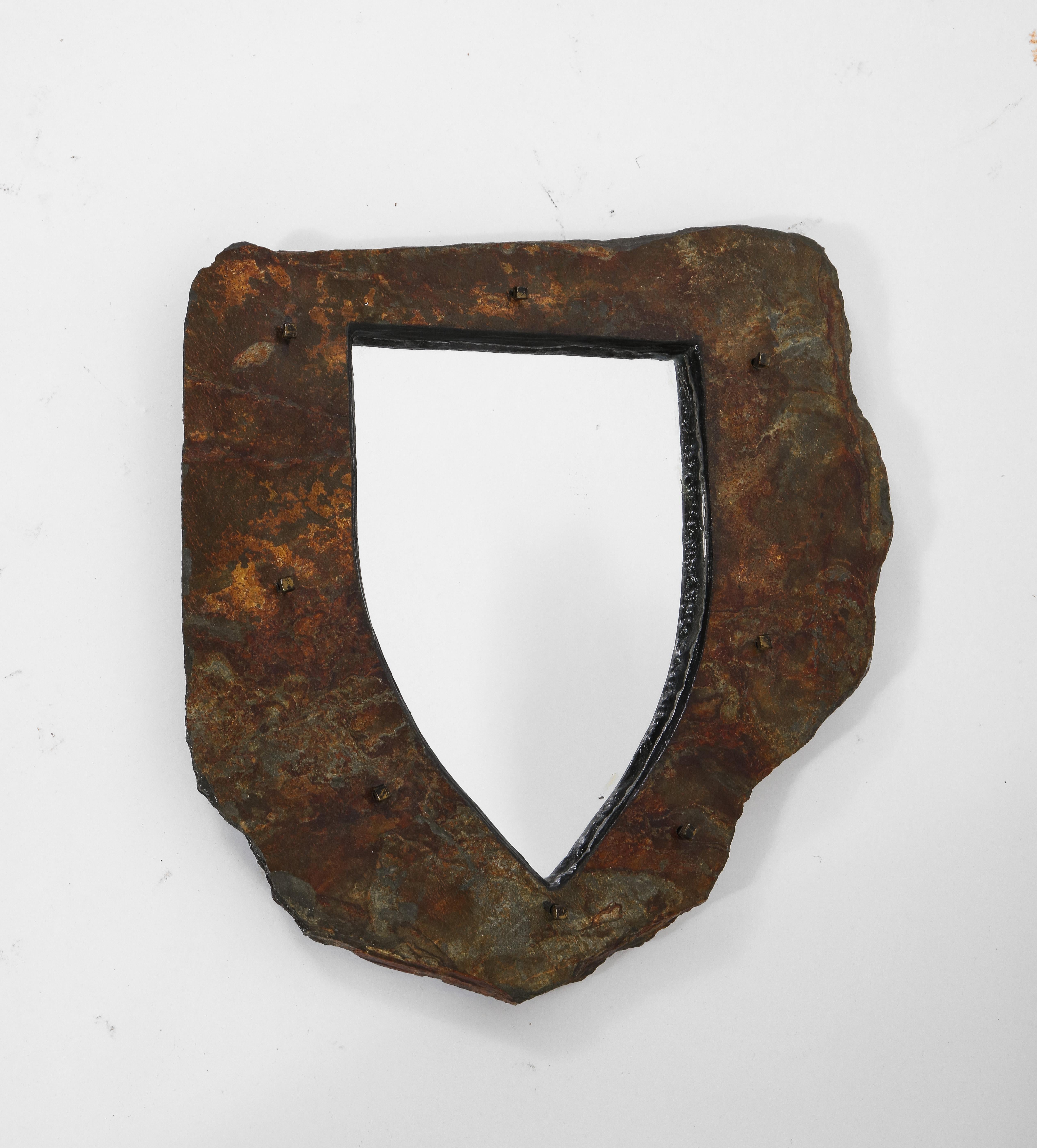 This distinct shield-shaped Brutalist mirror is composed of raw-edged schist in a deep coffee hinted with ochre. Metal rivets punctuate the frame of rough stone.