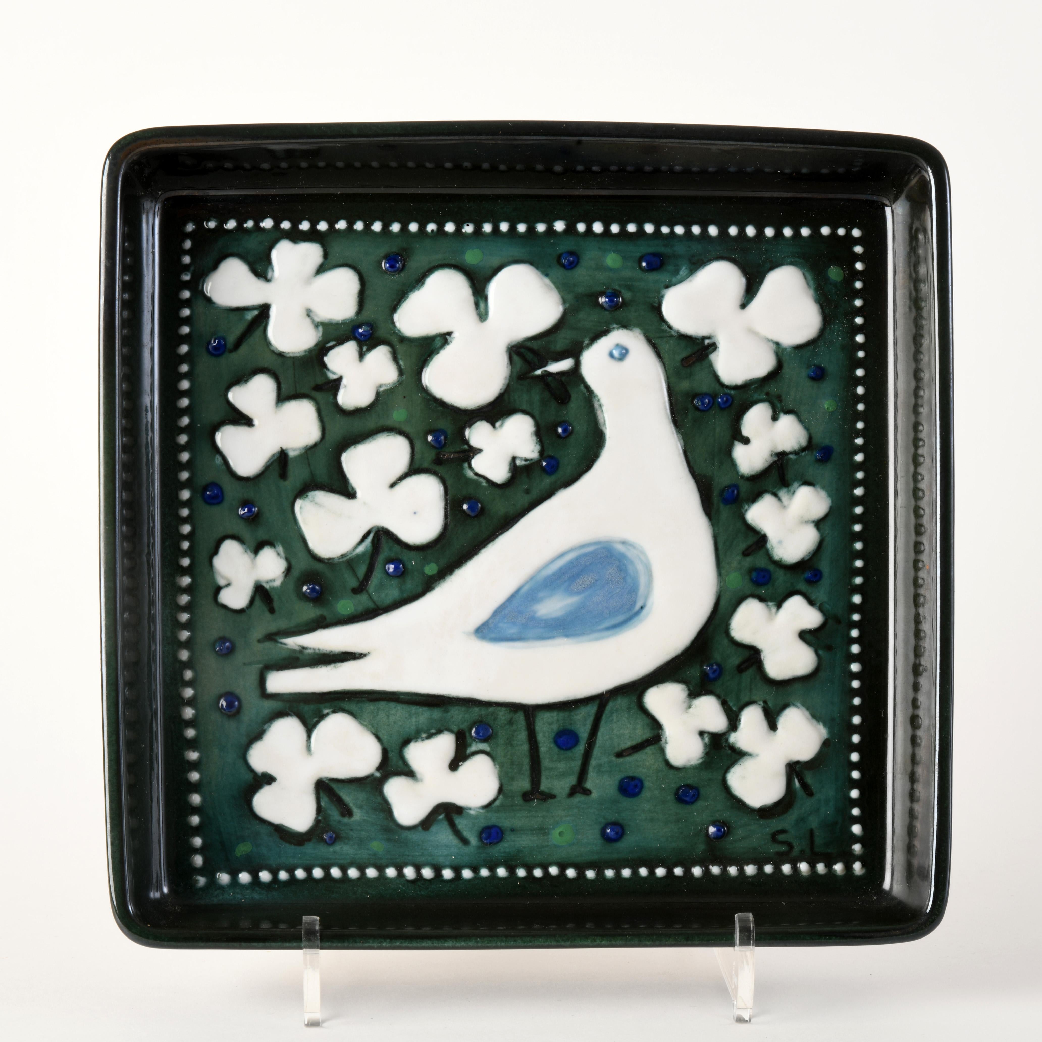 Small square dish finely enamelled in relief by Swedish artist Sylvia Leuchovius (1915-2003) in the 1960s/70s. The decoration depicts a white bird in the centre surrounded by white flowers on a dark green background dotted with blue spots. The edges
