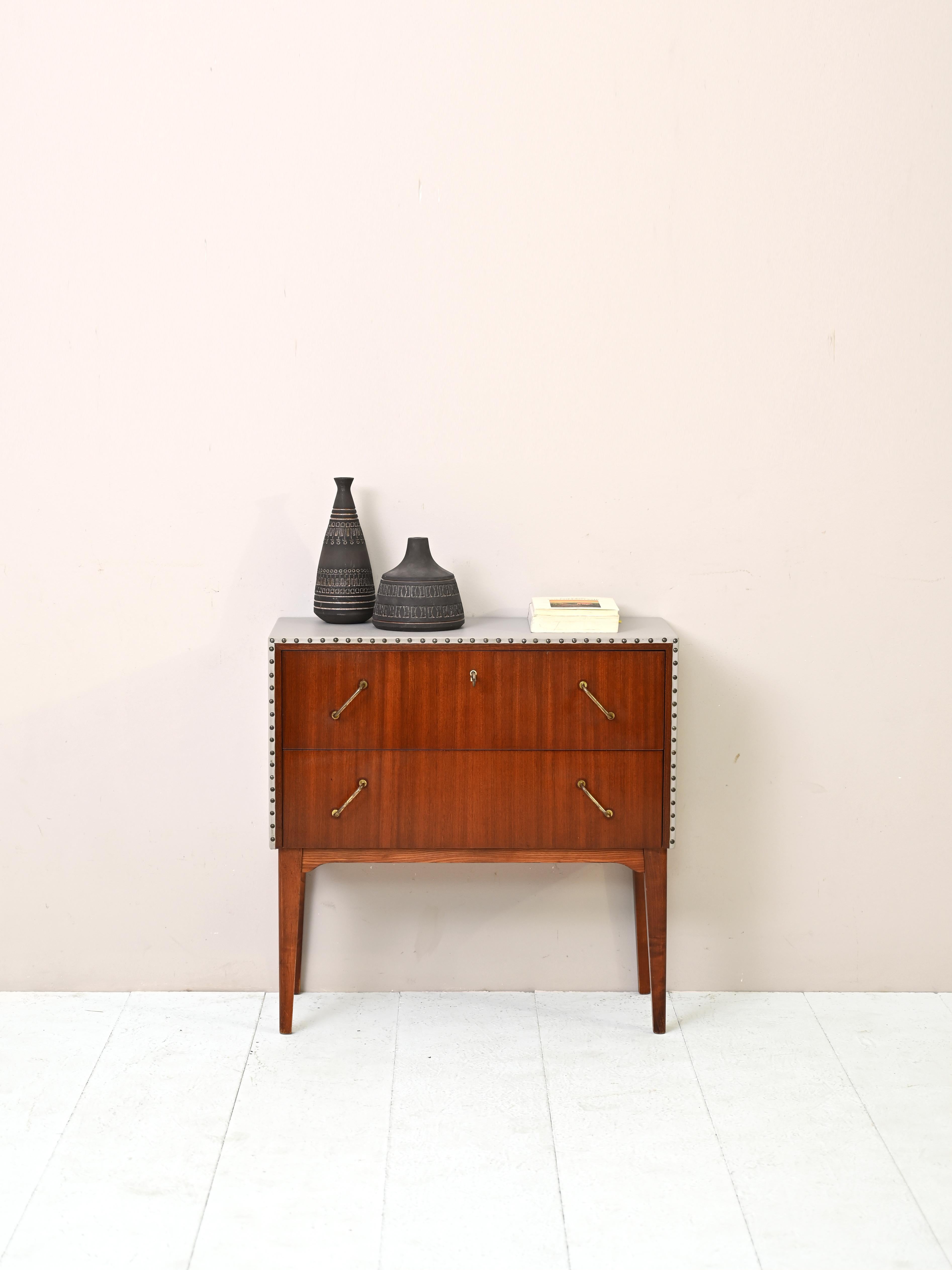 Small chest of drawers/bedside table of Nordic manufacture produced in the 1950s.

The entryway cabinet features cream-colored leatherette upholstery on the top and sides. The small metal studs drawing the front edge recall the handles.

An