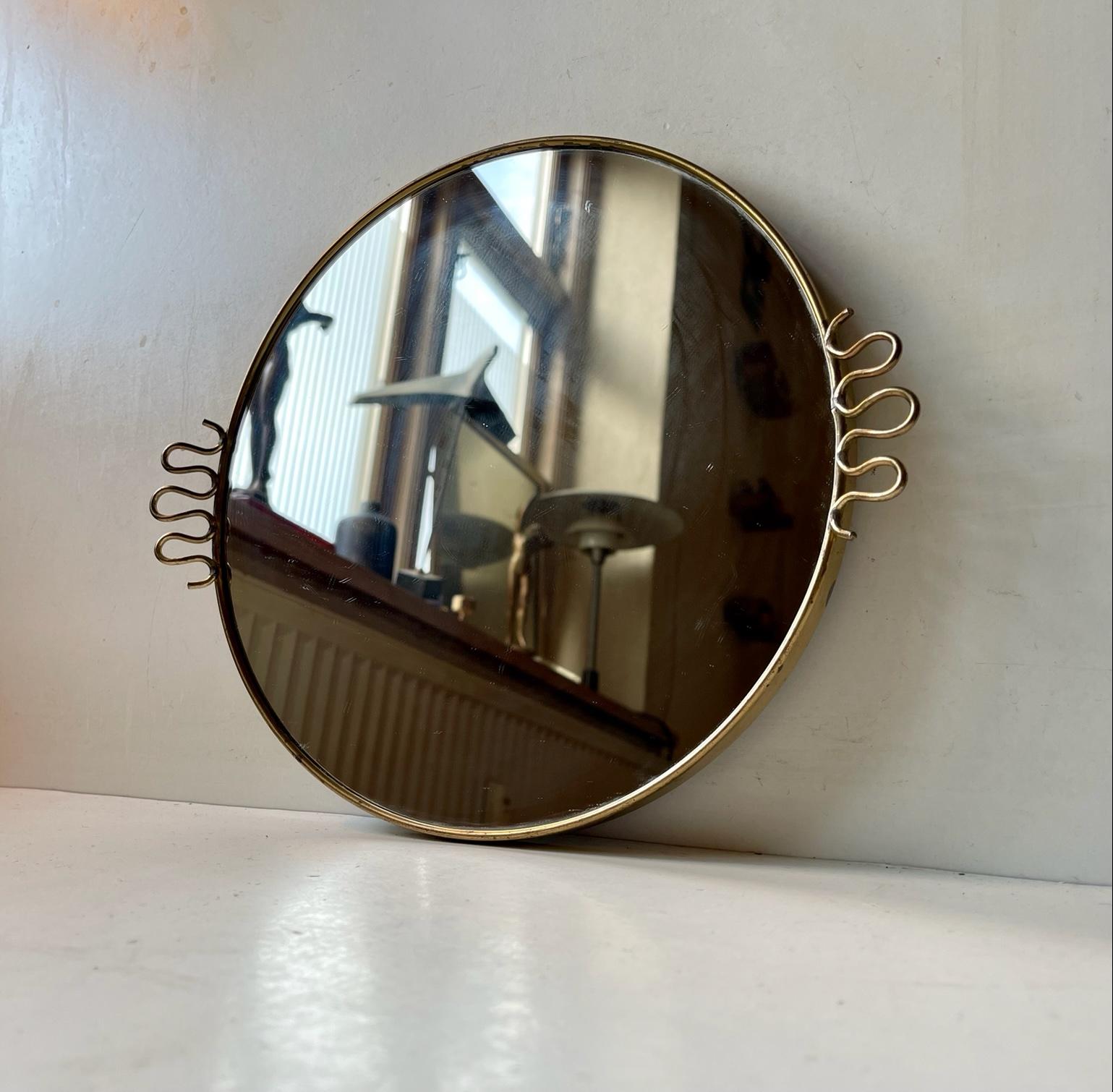 A small wall hung brass mirror. Suitable as vanity/make-up mirror. It features stylish brass loops reminiscent of those seen on Josef Franks Mirrors for Svensk Tenn. Measurements: D: 26 cm, Dept: 2.5 cm. The mirror glass and back-plate has been