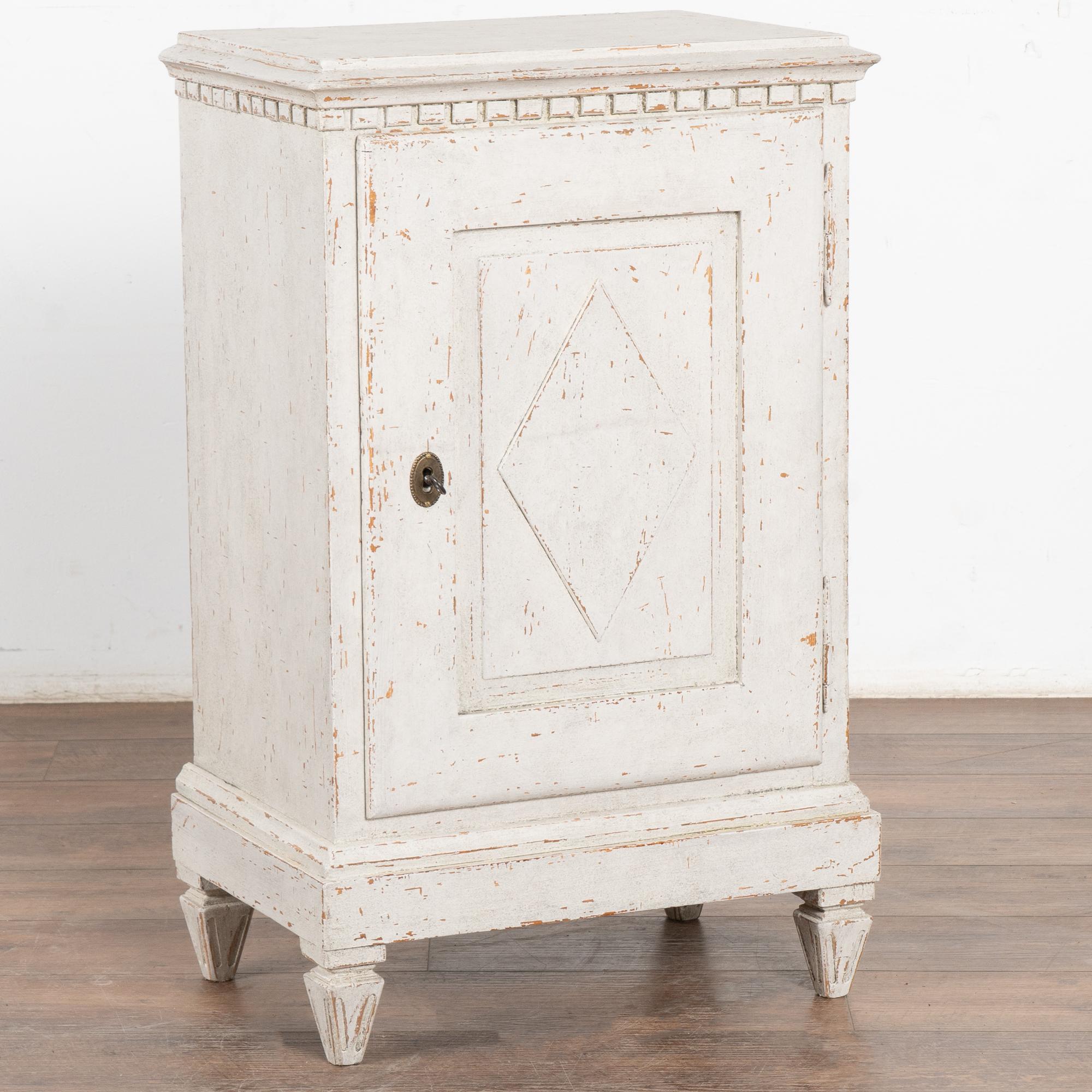 Small European country white painted nightstand or side table with dentil molding standing on fluted tapered feet.
Traditional diamond motif on single panel door which opens to reveal interior shelves.
Newer professionally applied antique white