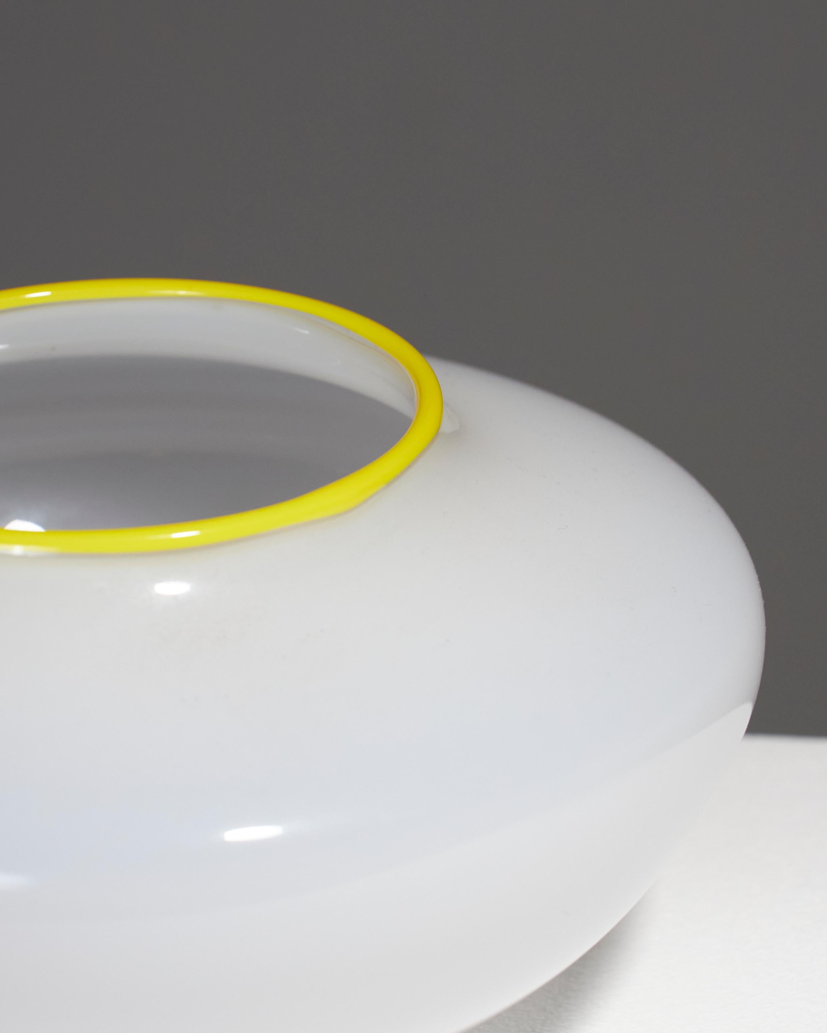 Vaso a corpo rotondo in vetro bianco opaco, realizzato negli anni '60. Il collo è evidenziato da una banda gialla. Poggia su una piccola base circolare. In ottime condizioni