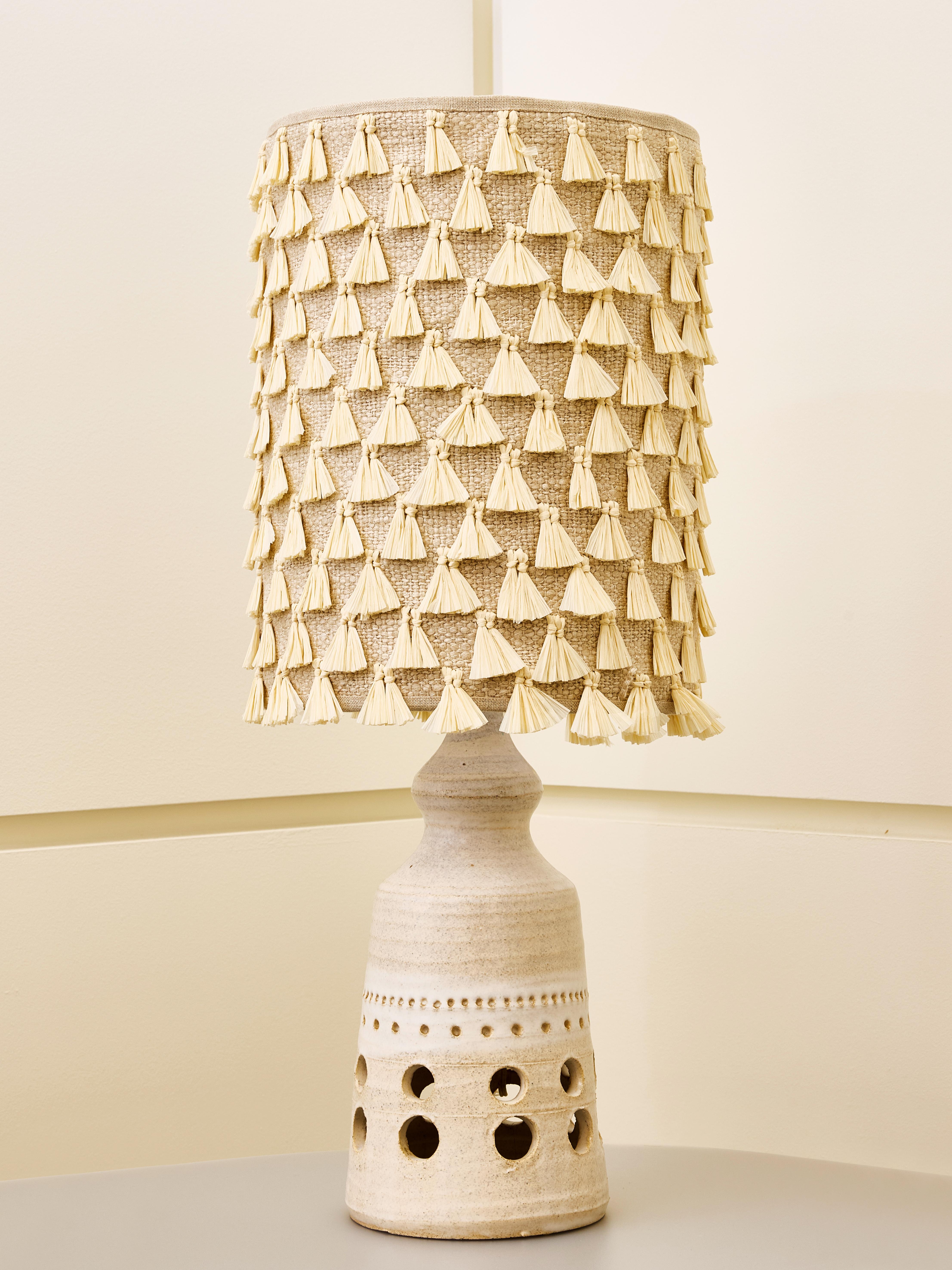 Cette lampe présente une esthétique chaleureuse et artisanale qui mêle matériaux naturels et design artistique. La base est en céramique émaillée blanc cassé, présentant des découpes circulaires et de délicats détails en pointillés qui lui donnent