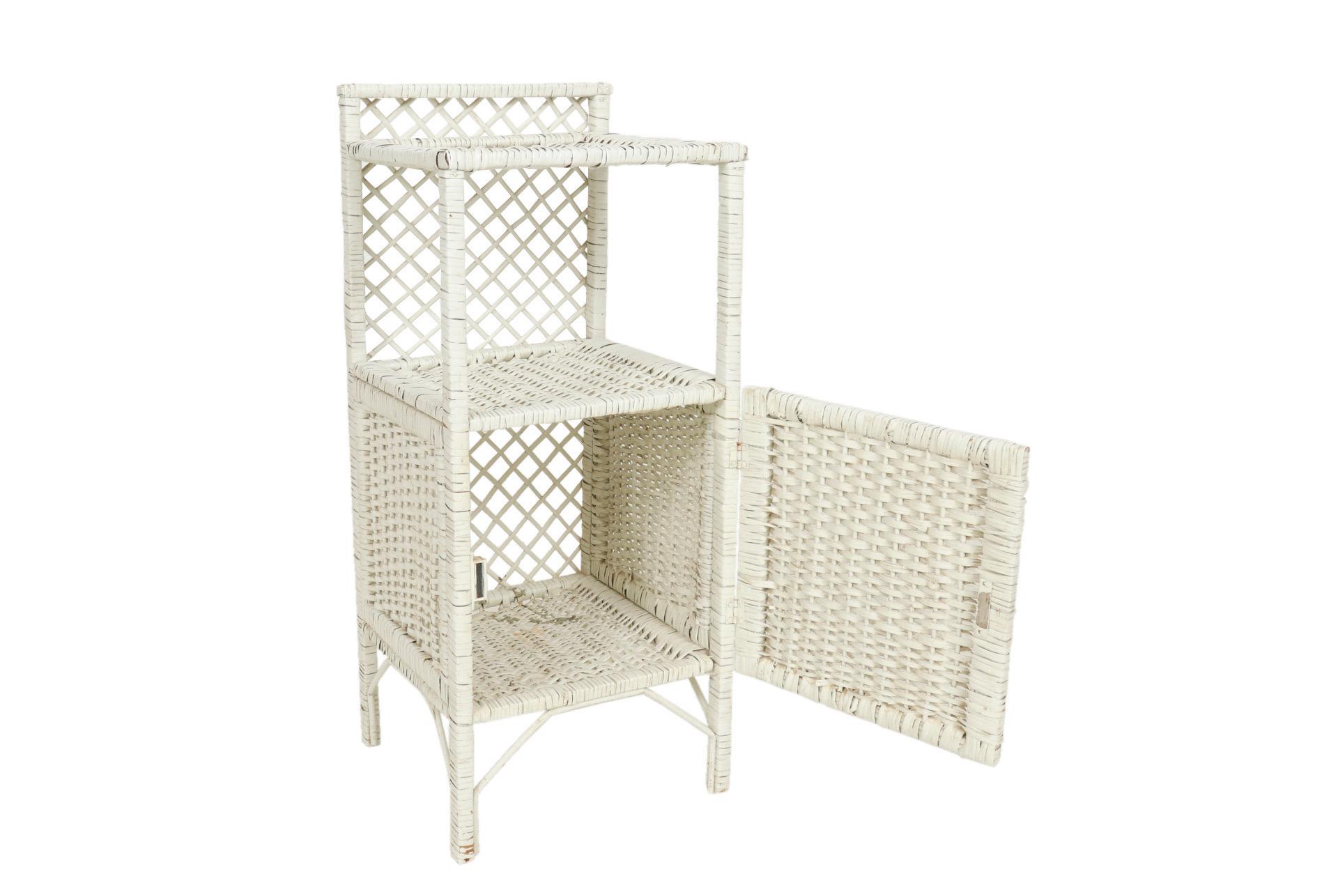 A small white cabinet made of wicker woven rattan. A single cabinet opens with a round wooden bead handle. Above is a single square shelf. The back is decorated with an open lattice weave.
 